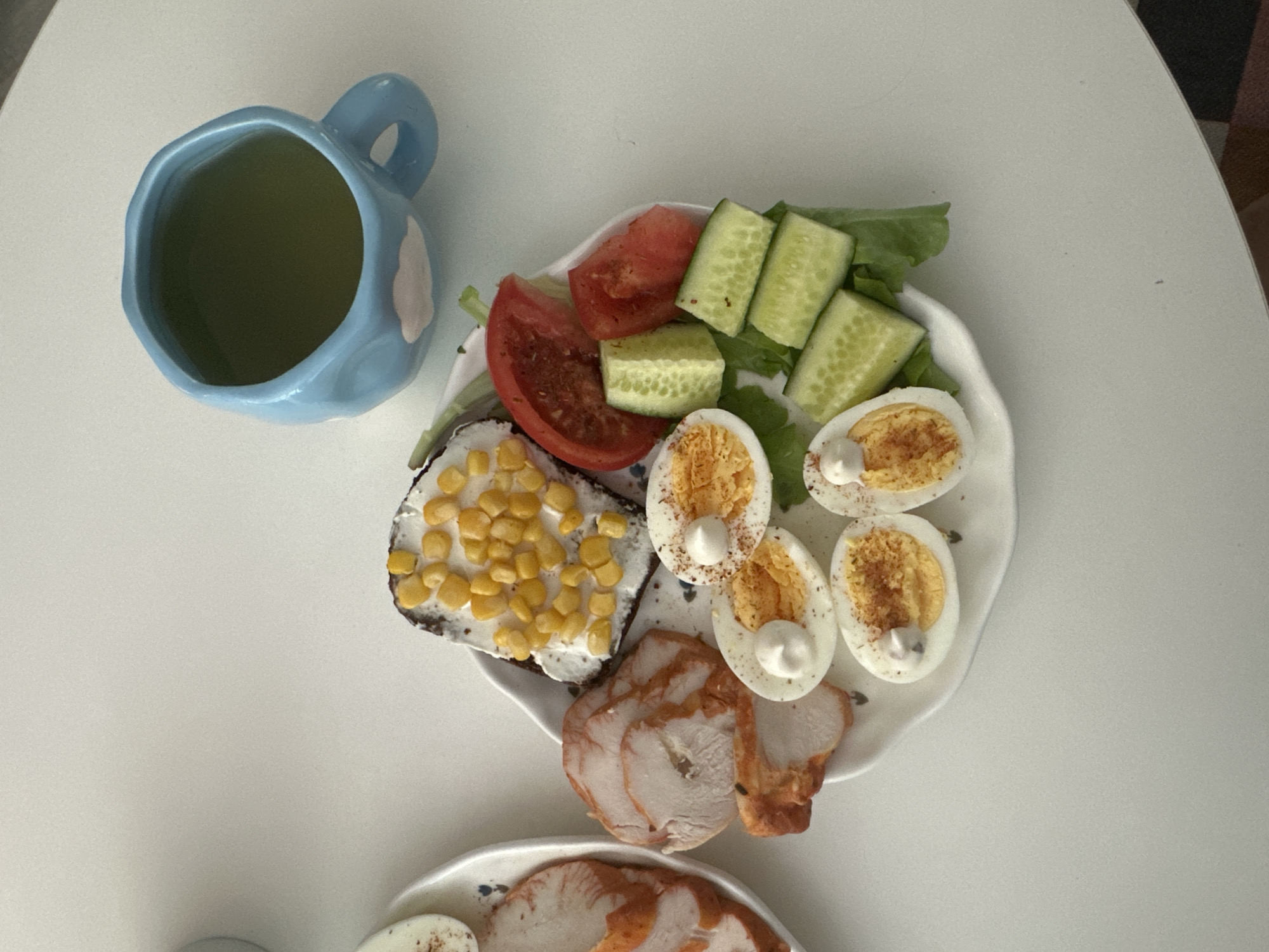 A high-angle, overhead shot shows a breakfast spread on a white, round table. In the upper left, a light blue ceramic mug with a cloud pattern holds a green liquid, likely tea or juice. To the right of the mug is a white, irregularly shaped plate containing a meal. The plate is divided into sections. The top section holds a salad of sliced tomatoes, cucumber rounds, and leafy greens. Below this, to the left, is a slice of dark bread topped with a white spread and corn kernels. To the right of the bread are several halves of hard-boiled eggs, each garnished with a dollop of white sauce and sprinkled with paprika or a similar spice. Below the eggs are several slices of cooked turkey breast. Another white plate with more sliced turkey breast is visible in the lower part of the frame. The overall scene suggests a simple, healthy breakfast. The lighting is bright, indicating it could be daytime. There is no visible text in the image. The setting appears to be domestic, possibly a kitchen or dining area.