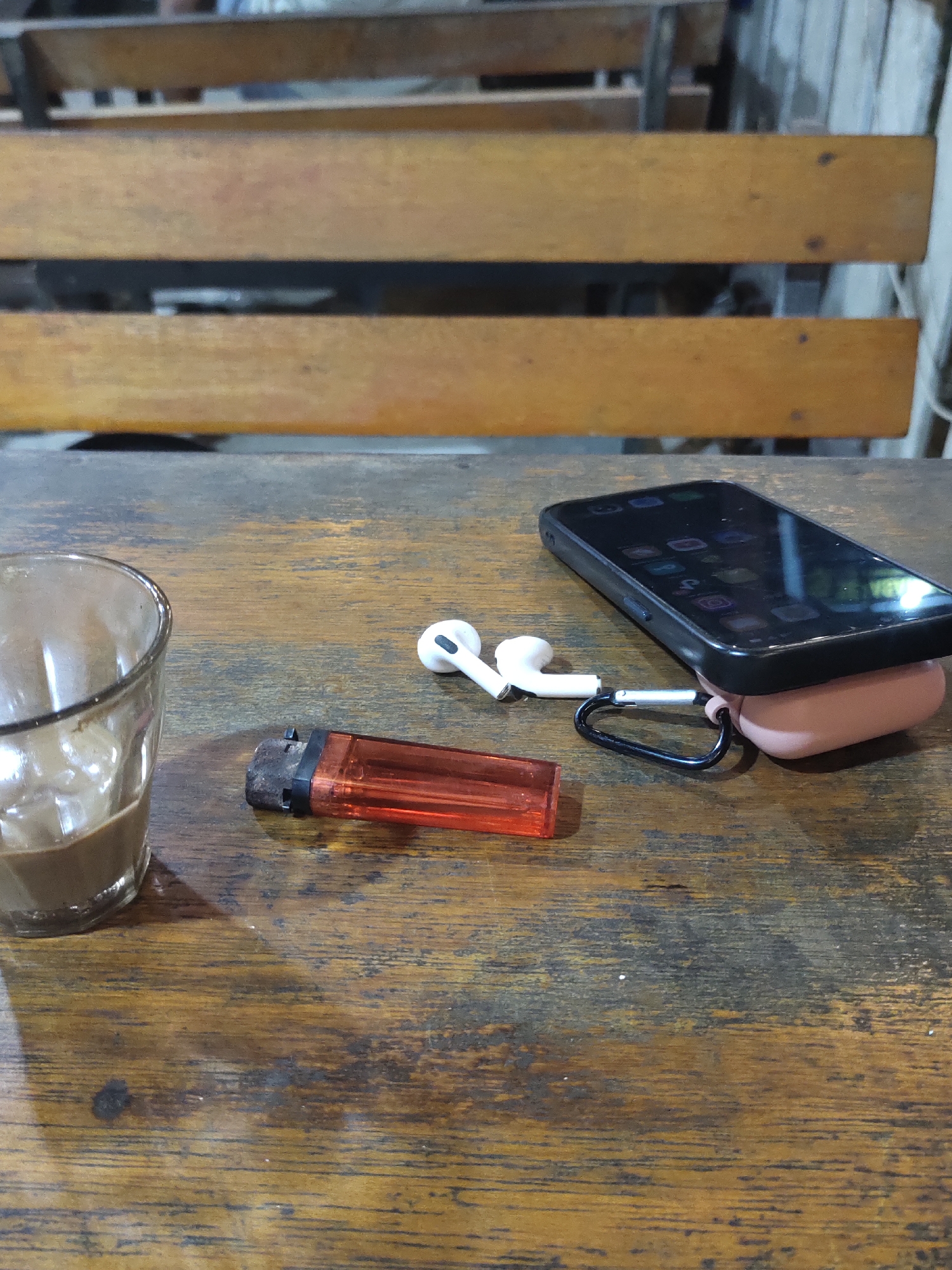The image depicts a close-up, eye-level view of a wooden table with several objects. On the left, a clear glass contains a light brown liquid, likely a beverage with ice. To the right of the glass, a red disposable lighter lies horizontally. Further to the right, a pair of white earbuds is connected to a pink charging case, which is partially underneath a black smartphone. The smartphone's screen displays various app icons. The background consists of wooden benches, suggesting an outdoor seating area or a cafe. The lighting is dim, with some reflections visible on the phone screen. The scene is situated in Keudematangkuli, Indonesia.