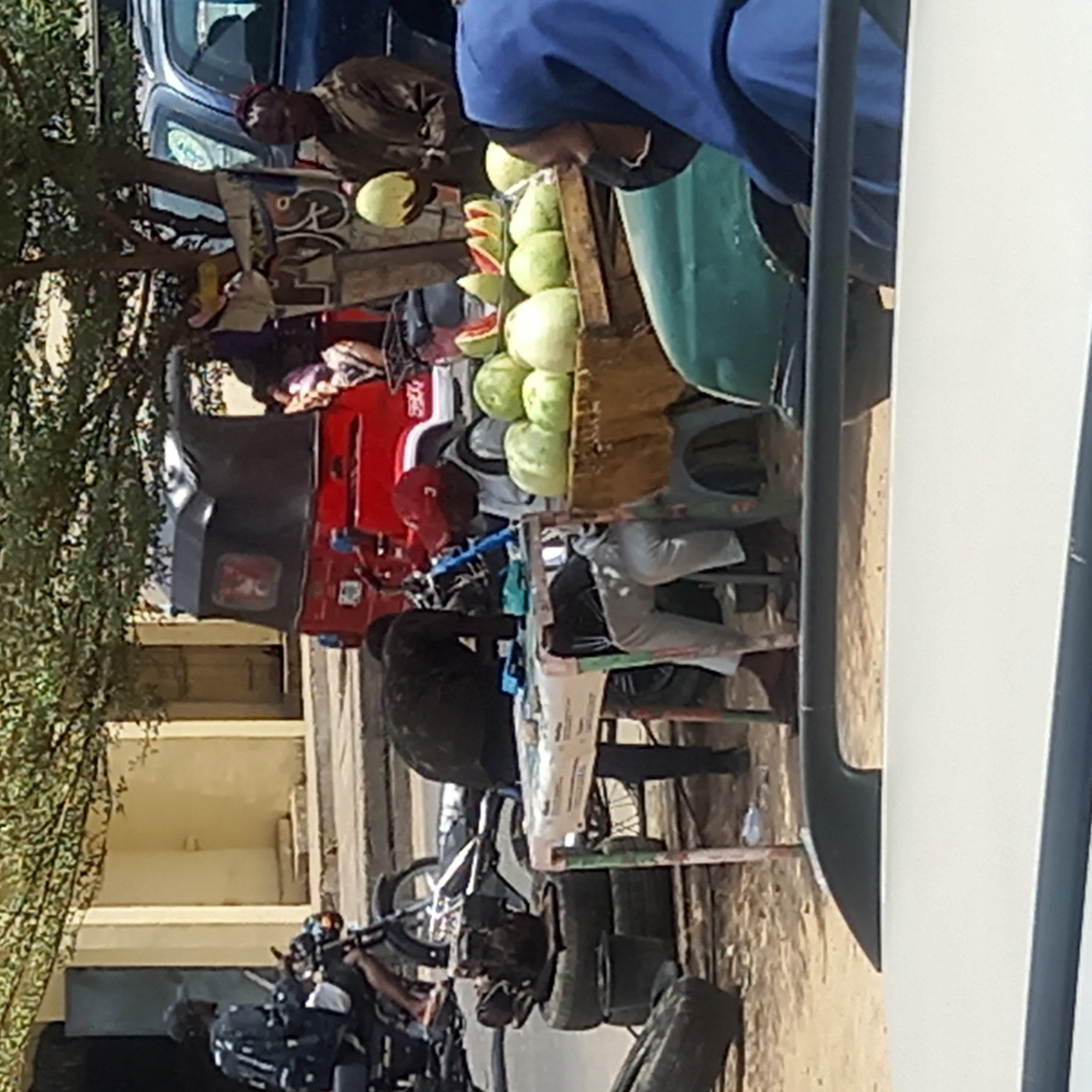The image shows a street scene in Dadi Riba, Nigeria. A man stands behind a cart filled with watermelons, some whole and some cut open to display the red flesh. There's a motorcycle repair shop next to the watermelon stand. A "keke napep" (motorized tricycle taxi) is parked nearby. Other parked motorcycles line the street, with people visible, some working on the motorcycles. The scene seems to be in a commercial area, in front of a single-story building.