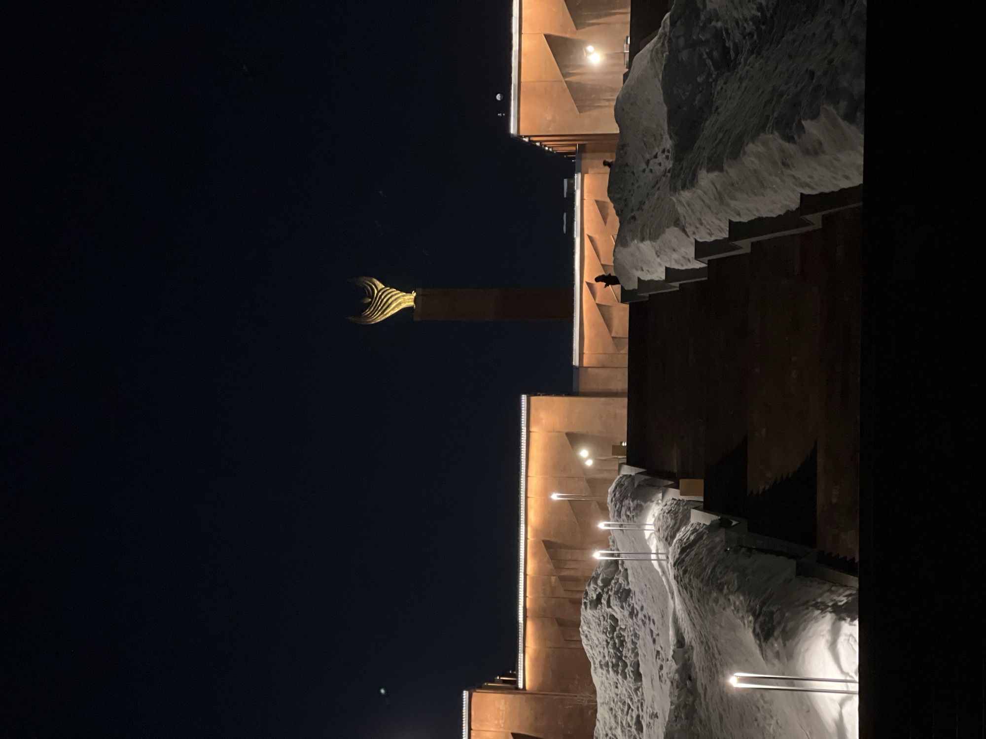This image captures a nighttime winter scene at the Monument to Kul Gali (also known as Winged Bars) near the Kazan Kremlin Museum-Reserve in Kazan, Russia. A tall, slender, reddish-brown column topped with a brightly illuminated, golden, abstract winged sculpture dominates the middle ground against a dark night sky. Flanking the monument are two large, reddish-brown buildings with flat roofs, each featuring horizontal strips of bright white perimeter lighting along the cornice and additional upward-facing facade lights. The lower sections of these buildings exhibit angular architectural elements. In the foreground, a wide, dark, ascending staircase leads towards the structures. Large, irregular mounds of white snow are accumulated on either side of the staircase. Several vertical, slender, cylindrical lights emitting a bright white glow are embedded within these snowdrifts. Two dark, indistinct human silhouettes are visible on an elevated platform in front of the reddish-brown buildings: one to the left of the monument's base and another further to the right, partially obscured.
