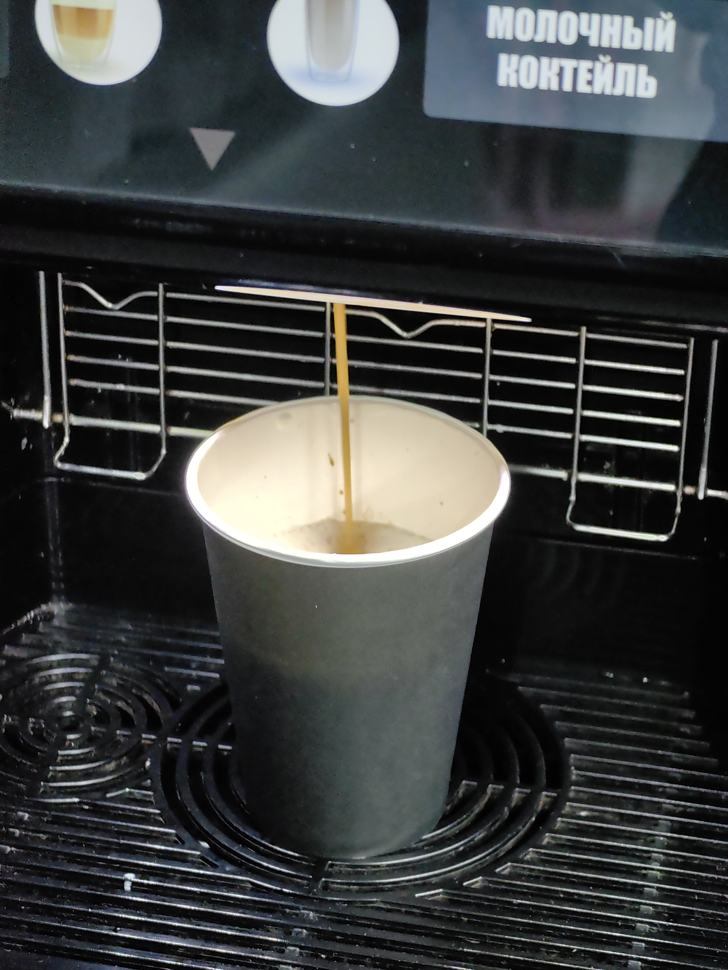 An automatic coffee machine dispenses a beverage into a gray paper cup. The cup is positioned on a black, gridded surface. Above the cup, a thin stream of light brown liquid is pouring from a nozzle. In the background, a screen displays white Cyrillic text that reads "МОЛОЧНЫЙ КОКТЕЙЛЬ" (milk cocktail). Two circular icons representing drinks are also visible on the screen. The overall environment appears to be indoors, likely in a public establishment. The image was taken in Minsk, Belarus.