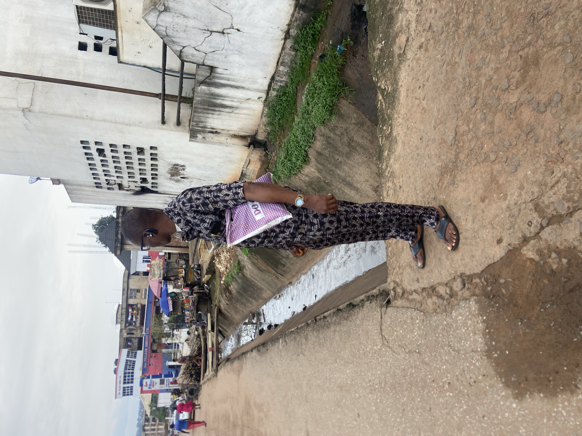 A man wearing a black and white patterned outfit and dark sandals stands on a paved walkway in Nkpor, Nigeria. He is seen from the waist down, with a purple and white folder in his left arm. He wears a wristwatch on his left wrist and earbuds in both ears. Behind him, a concrete drainage ditch runs alongside the walkway. The ditch contains murky water and some debris. The walkway is cracked and uneven, with puddles of water in places. In the background, there are buildings, including a multi-story structure with signs for "PILLARS ENERGY LTD." and "DO." People can be seen walking on the road in the distance, and there are market stalls with umbrellas and various items for sale. The sky is overcast.