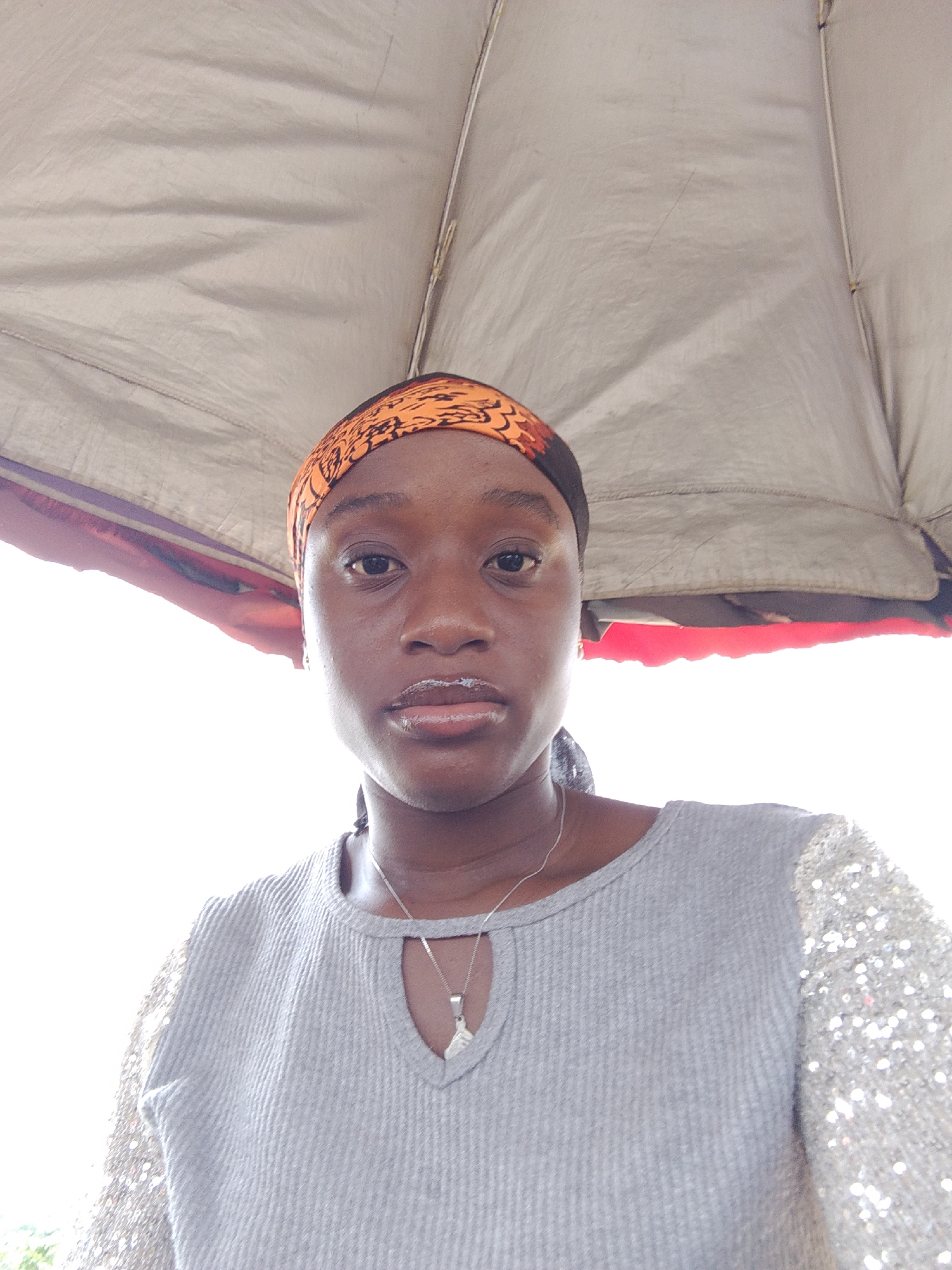 A young Black woman is centered in the frame, wearing an orange patterned headscarf and a gray top with sequined sleeves. She has a silver necklace with a pendant. The woman stands beneath a tent or awning, judging by the fabric visible overhead and to the sides. The background is brightly lit, possibly indicating daylight. The location is potentially in Umuahia, Nigeria, as specified in the prompt. The absence of additional visual information makes it impossible to confirm this location or to elaborate on any specific actions, interactions, or objects within a broader environmental context.