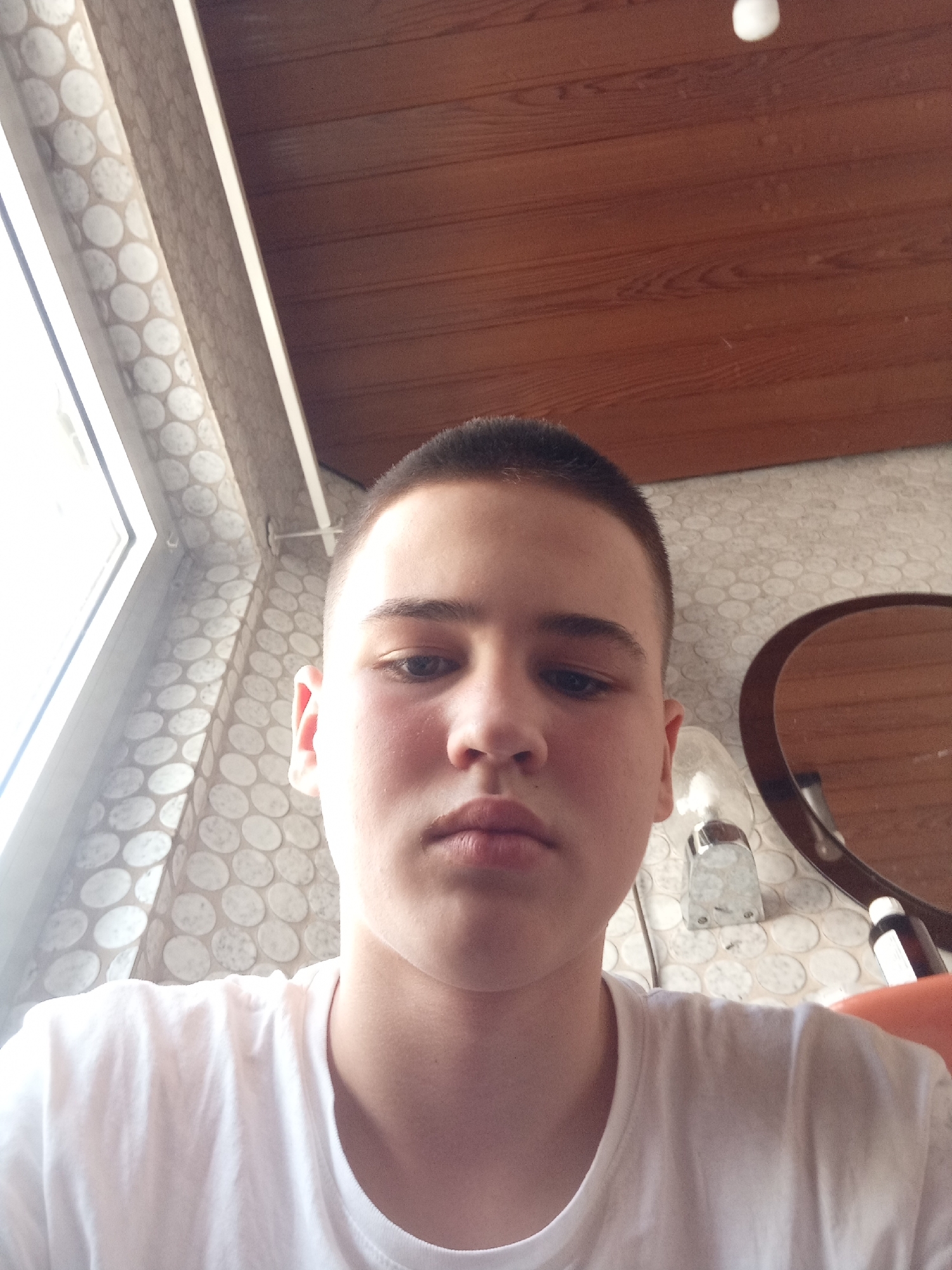 A young male with short, dark hair and fair skin is looking at the camera, taking a selfie. He is wearing a white t-shirt. The background consists of a patterned wall, a wooden ceiling, and a window with a patterned shade. A round mirror with a dark frame is visible to the right of the subject. There is also a dispenser-like object and a bottle in the background. The lighting suggests an indoor setting. The image was taken at an unknown location in Kaiserslautern, Germany.