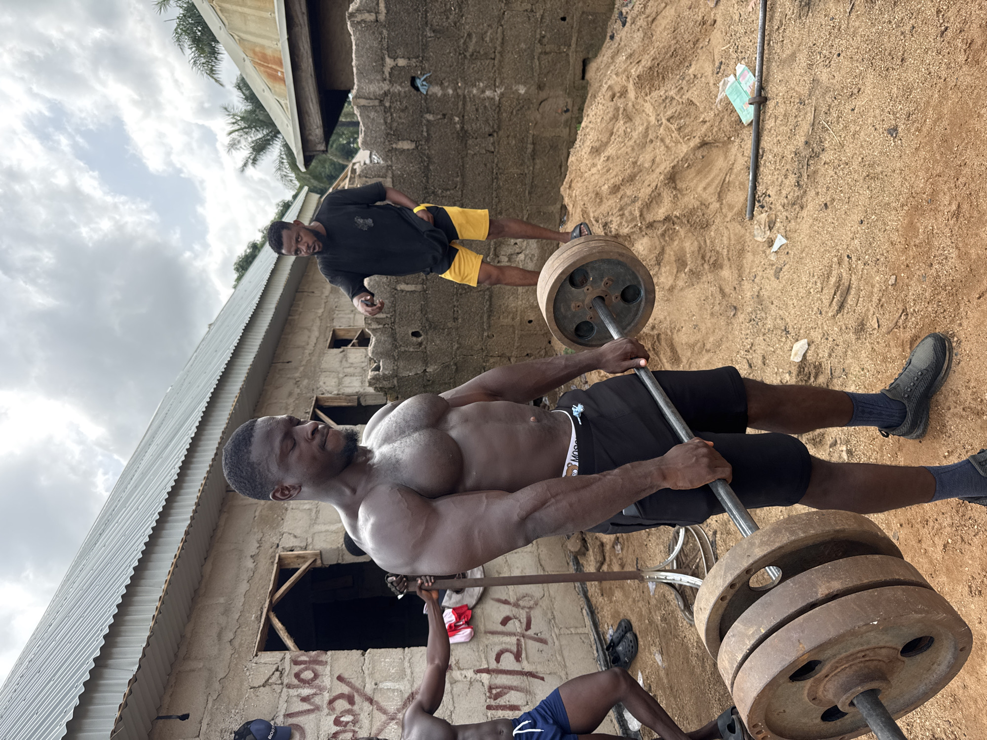 The image depicts an outdoor weightlifting scene, likely in a residential area, possibly near Akwanga, Nigeria. A muscular, shirtless man is in the foreground, in the midst of lifting a barbell loaded with multiple weight plates. He is wearing black shorts. Another man, wearing a black shirt and yellow shorts, stands in the background, appearing to observe or spot the weightlifter. A third individual is partially visible in the lower left corner. The backdrop consists of a roughly constructed building with an unpainted exterior and visible graffiti. The ground is sandy. The sky is overcast.