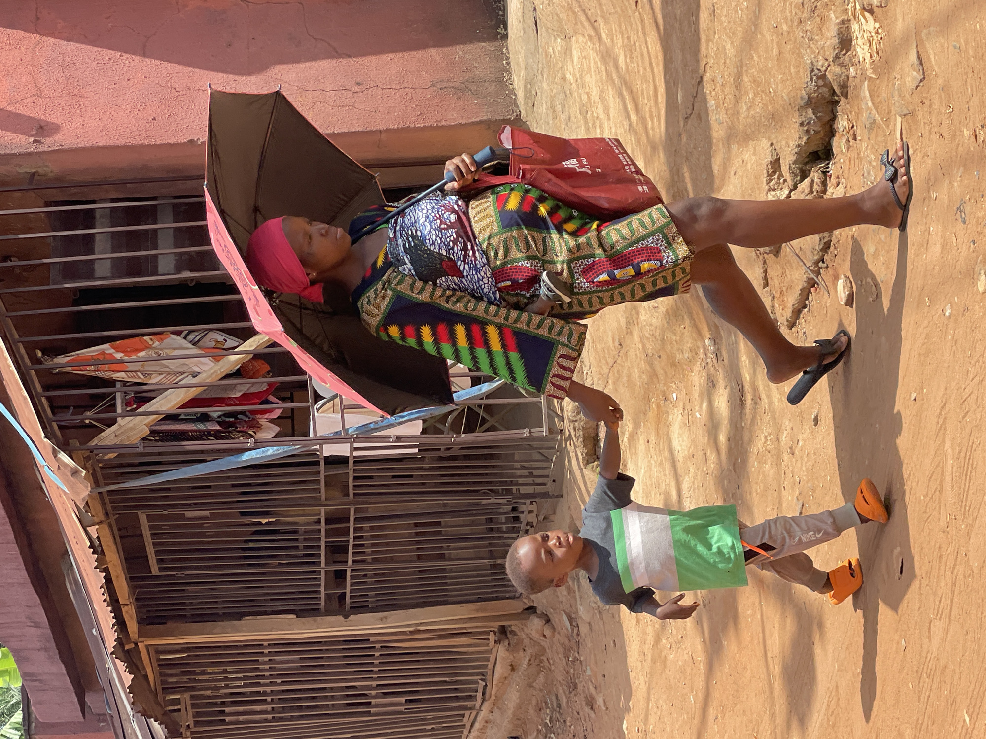 A woman and a child are walking on a dirt road next to a building in Onitsha, Nigeria. The woman is wearing a colorful dress and black sandals. She is holding an umbrella over her head and carrying a red shopping bag. The child is wearing a gray t-shirt with a green and white stripe, gray sweatpants, and bright orange clogs. He is holding the woman's hand. The building is made of red brick and has a corrugated metal roof. There are clothes hanging on a line outside the building.