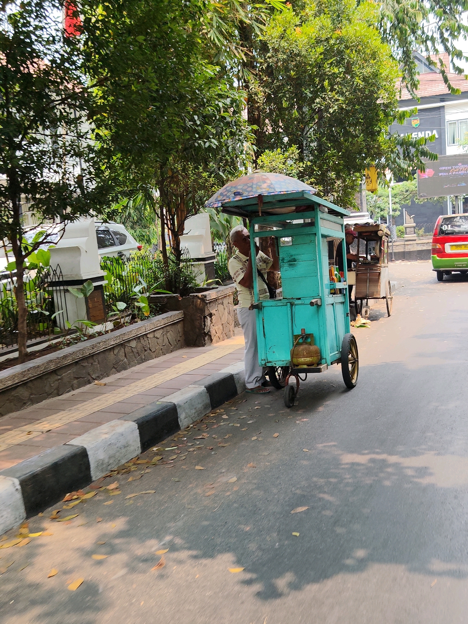 In Purwakarta, Indonesia, on a paved road, a man with gray hair stands behind a teal-colored food cart. The cart has a patterned umbrella and a propane tank attached to its side. In the background, a second food cart is visible, and a red and green vehicle is driving on the road. The sidewalk features a black and white striped curb and textured paving. Trees and buildings line the street, suggesting an urban environment.