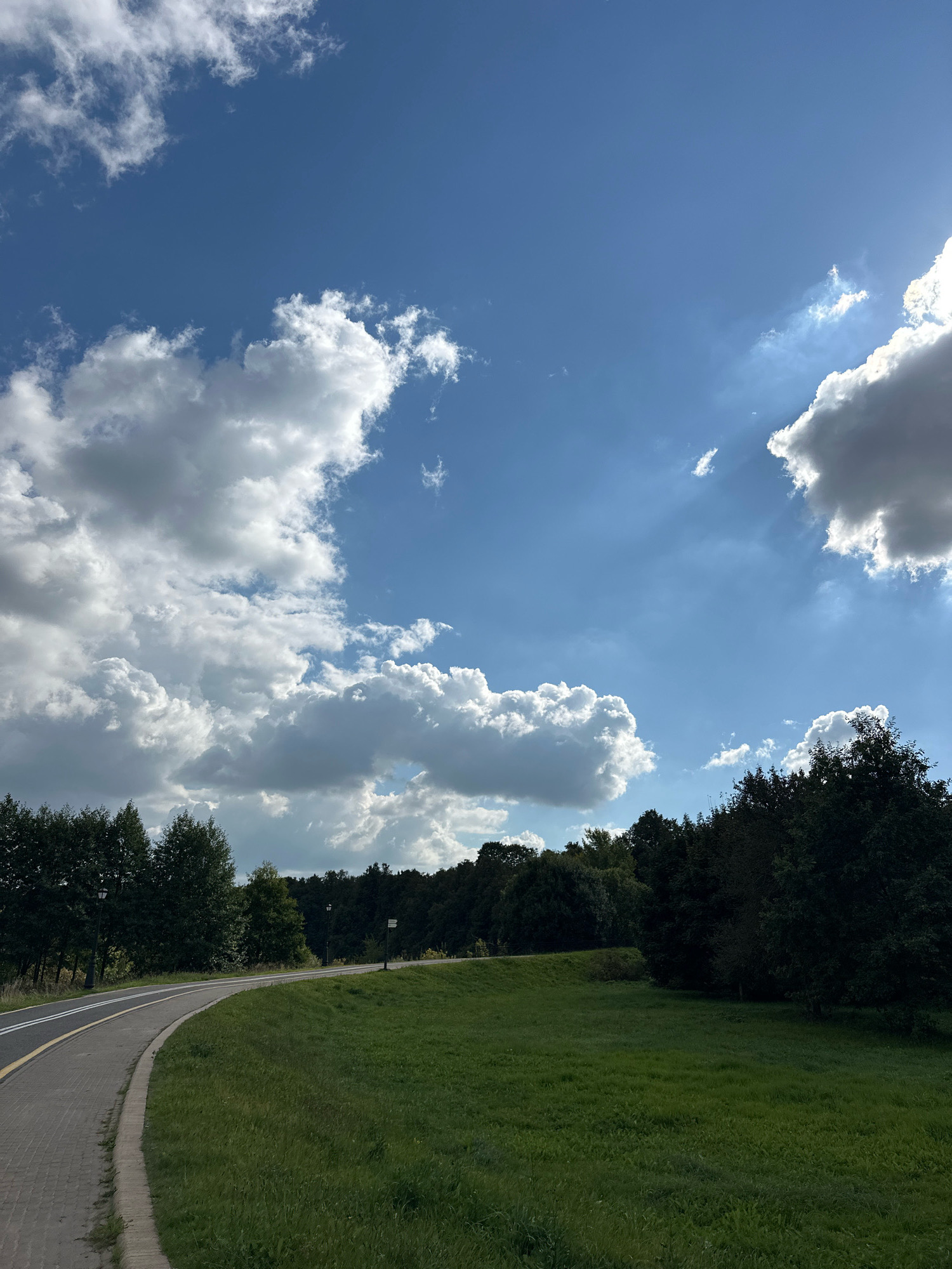 The image depicts a curved asphalt road and a brick pathway in a park-like setting. The road is bordered by a strip of yellow and white paint. To the right of the path, a lush green grassy slope rises and is interspersed with trees and bushes. Further in the background, a dense forest forms a dark green line across the horizon. Two streetlights are visible in the distance along the road. The sky above is a vibrant blue with scattered white and grey clouds, and sunlight streams through from the upper right corner. The overall impression is of a scenic outdoor location suitable for recreation. This appears to be the area of the Minsk Sea, a reservoir located on the Svislach River, northwest of Minsk, Belarus.
