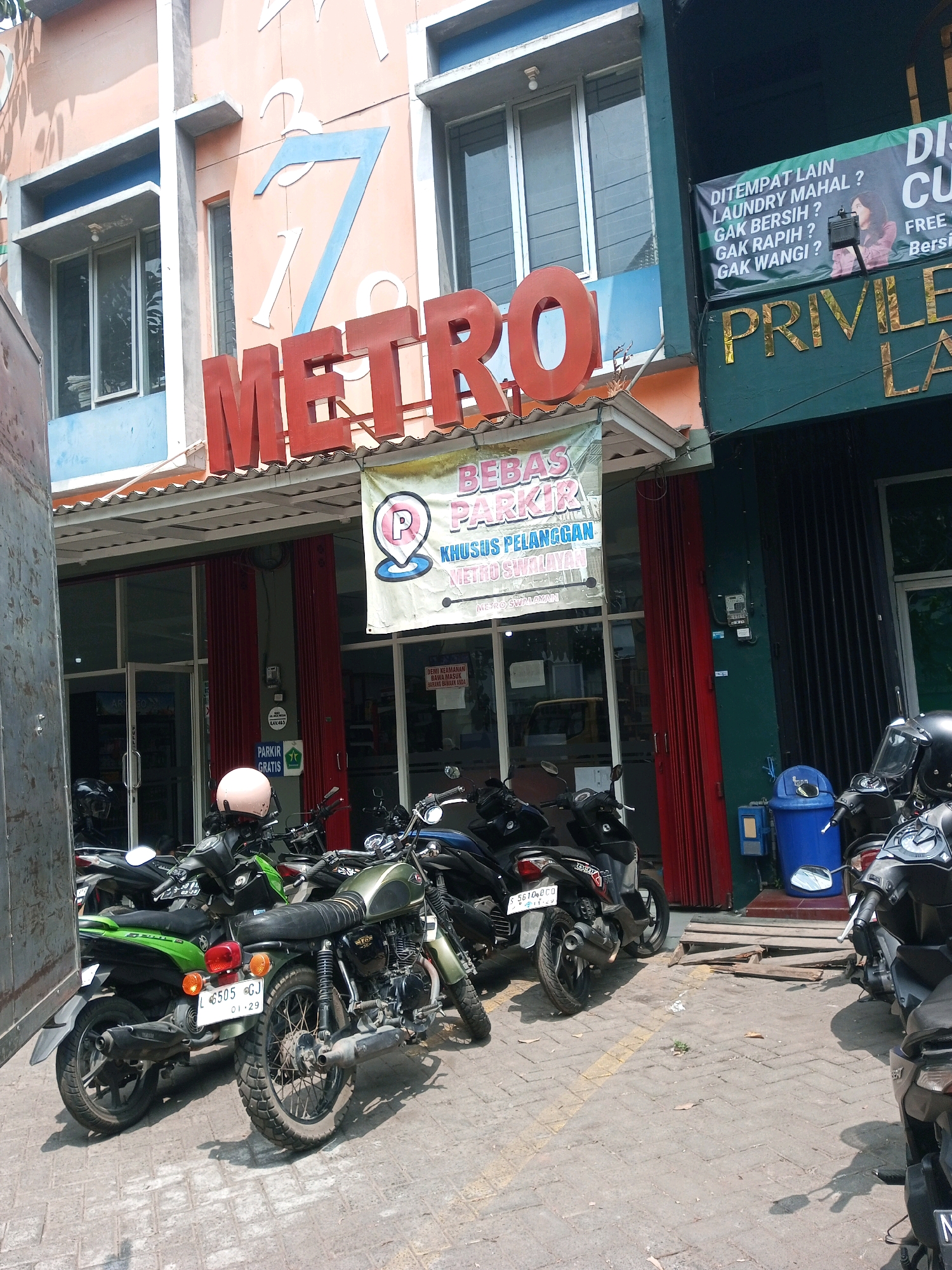 The image shows the exterior of a building in the city of Malang, Indonesia, with a prominent "METRO" sign in red letters. In front of the building, several motorcycles are parked. A banner with text in Indonesian, including "BEBAS PARKIR KHUSUS PELANGGAN" (Free Parking for Customers), is displayed. The building appears to be a commercial establishment. The environment is an outdoor urban setting with a paved ground.