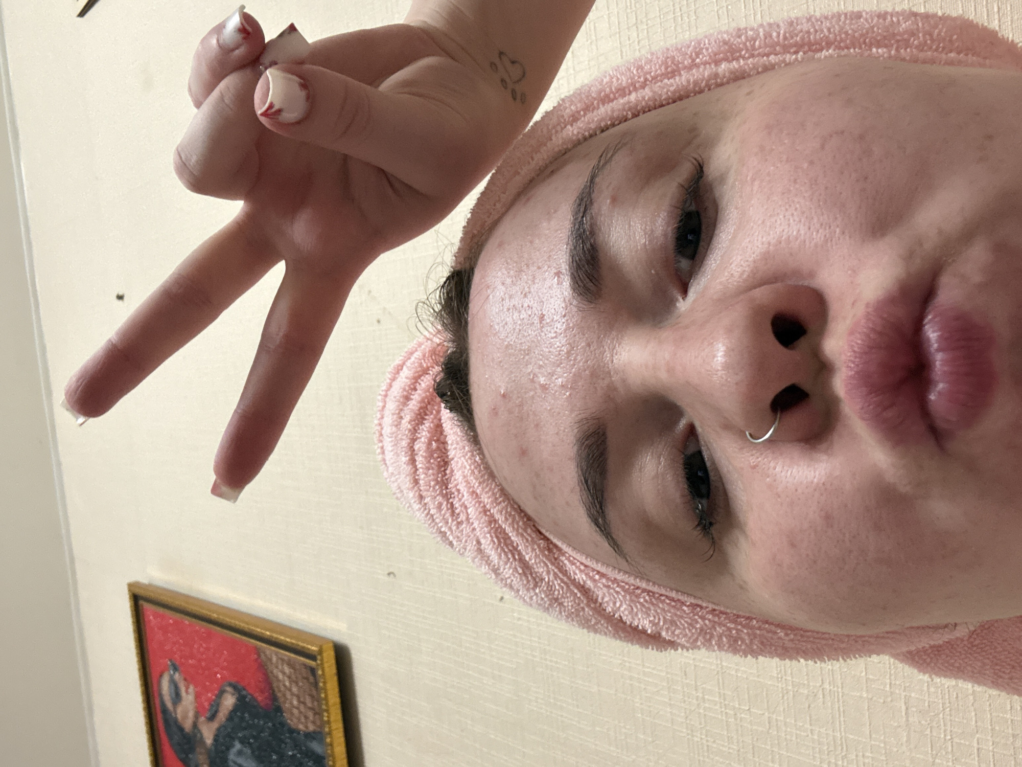A close-up, upside-down photograph depicts a person's face, wearing a pink towel around their head. They have fair skin, dark eyebrows, and a silver nose ring. Their lips are pursed. Above their head, a hand makes a peace sign. The fingers have white square tips with red designs. A tattoo of a paw print enclosed in a heart is visible on the wrist. In the background, a framed painting hangs on a light-colored wall. There is no evidence to confirm this is taken in Izhevsk, Russia.