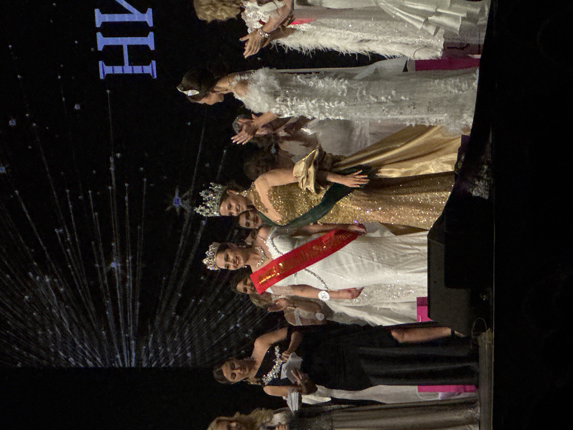 The image shows a group of women on a stage, possibly at a beauty pageant or similar event, in the city of Unknown, Unknown. They are dressed in gowns and some are wearing crowns and sashes. They are standing and appear to be waving or acknowledging an audience. There is a dark background with what seems to be a light design or projection. The lighting is focused on the stage. The general mood is celebratory.