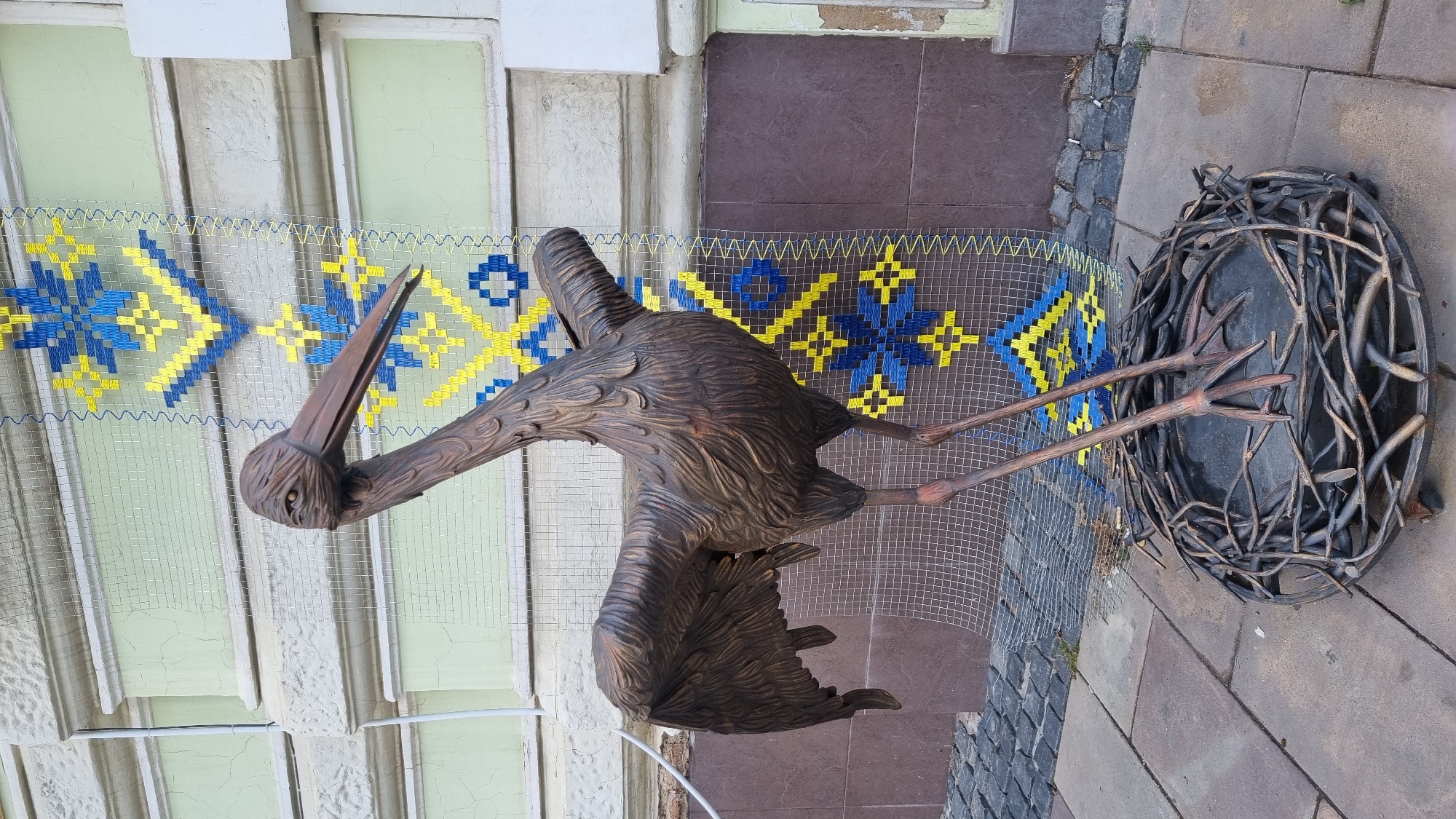 A bronze sculpture of a stork stands in a nest on a cobblestone sidewalk. The stork is depicted with its wings partially spread and its head turned to the left, with one eye visible. Behind the sculpture, a building with light green walls and decorative elements is visible. A large, rectangular mesh screen is hung on the building's facade, displaying a pattern of blue and yellow Ukrainian folk art motifs. The sidewalk is paved with light gray and dark gray rectangular stones, and the cobblestone area behind it is darker. The sculpture and its surroundings suggest an outdoor public space in the city of Chernivtsi, Ukraine, possibly near a historic building. There are no people or animals present in the image.