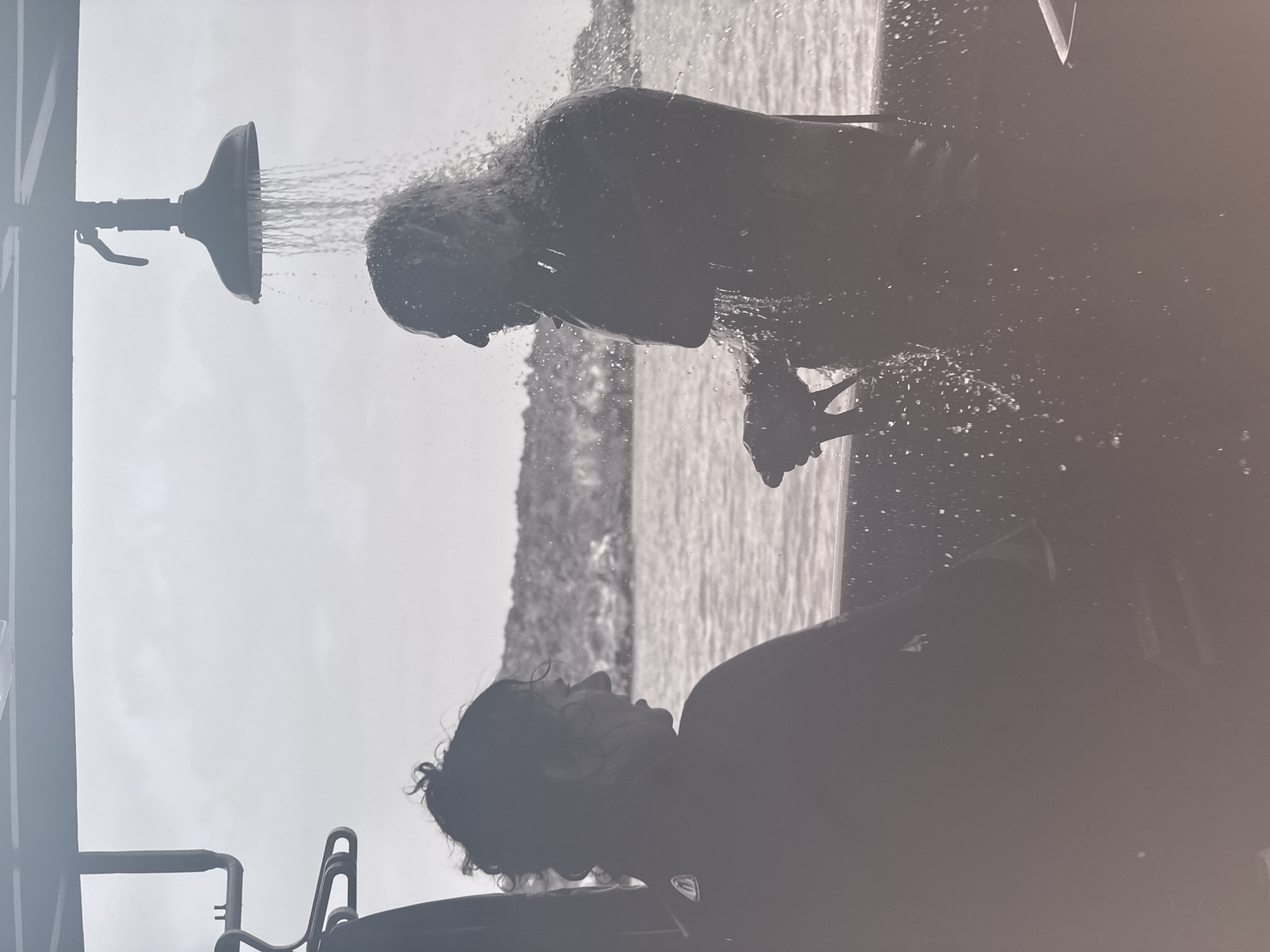 A monochromatic, low-angle shot depicts two individuals interacting with a shower fixture, possibly outdoors and near a body of water.
The first individual is shown directly under a showerhead, receiving a stream of water. They are silhouetted against a lighter background, making it difficult to discern specific features.
Below and slightly offset, a second person is positioned in the lower half of the image. Their face is tilted upwards, eyes seemingly closed or partially closed. Details of their features, such as hair and eyewear, are visible.
A shower fixture is prominently positioned on the left side of the image. It includes a showerhead dispensing water, a handle, and supporting framework.
The background is less defined, possibly showing the water and landscape.
Due to the monochrome nature of the image and lack of explicit landmarks, it is impossible to ascertain if the scene is set in Mexico City, Mexico.
