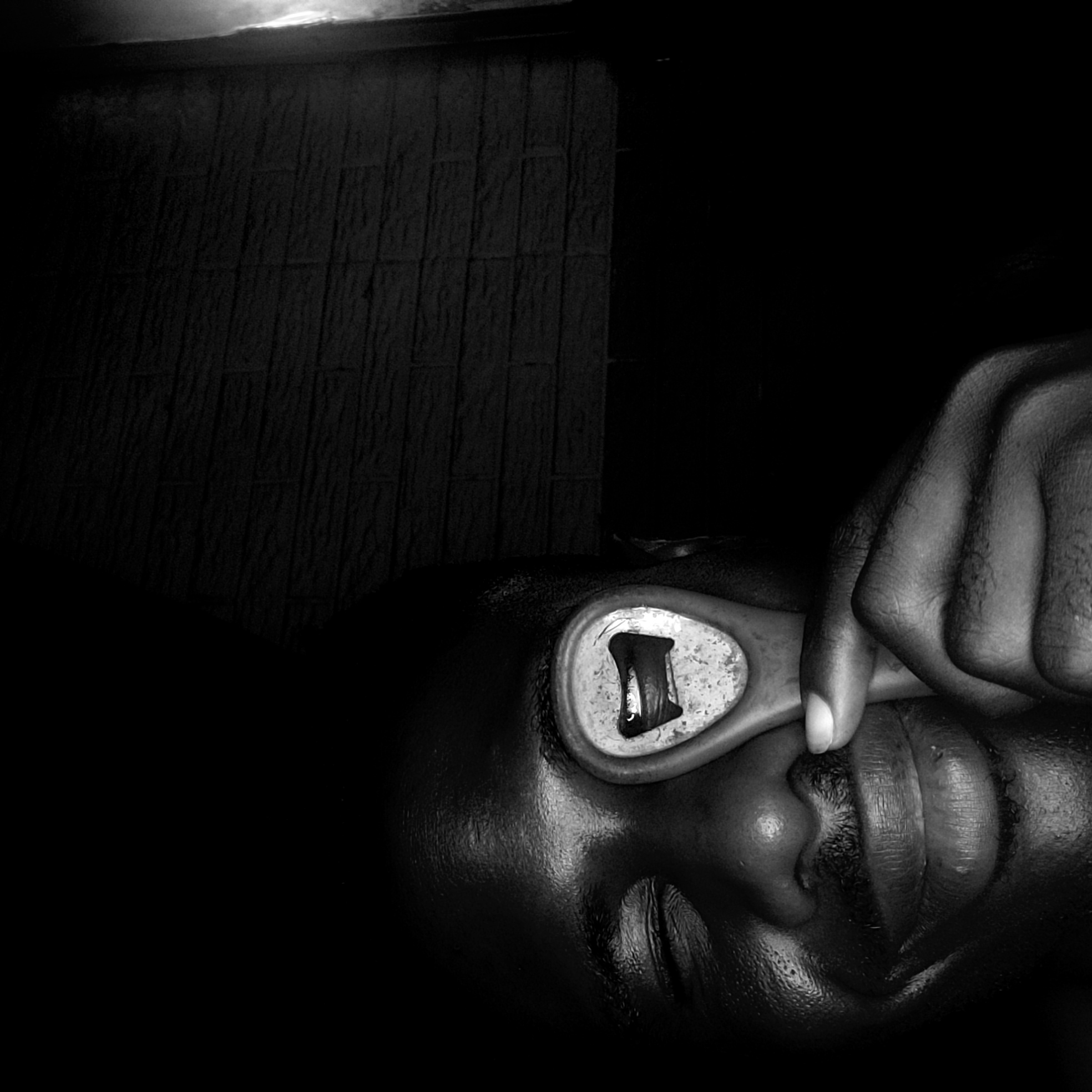 A close-up, low-angle, black and white image shows a person lying on their back. A bottle opener is placed over the person's eye. A hand holds the bottle opener in place. The setting appears to be indoors, with a wall featuring a brick-like pattern in the background. No indication of Narayi, Nigeria.