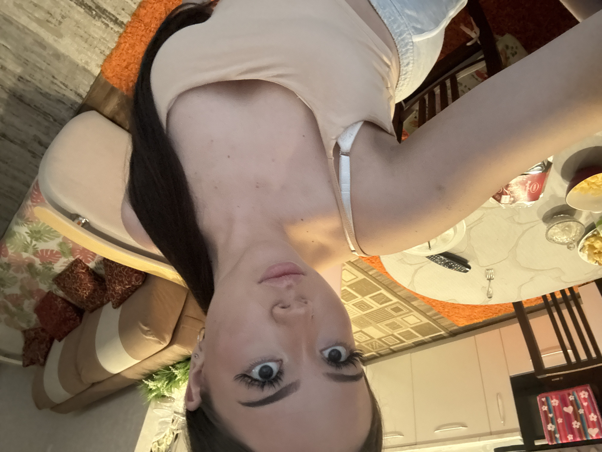 An upside-down selfie captures a young woman with long dark hair, wide eyes enhanced by dark eyeliner and long eyelashes, and slightly parted lips. She wears a light beige top with a visible white bra strap. The image is rotated 90 degrees counter-clockwise, giving the impression of her looking down at the camera from above.
The setting appears to be an indoor residence in Naberezhnye Chelny, Russia, likely a living or dining area with an adjacent kitchen. In the background, a light-colored sofa with striped and patterned pillows (featuring red and brown leaf motifs) is visible, alongside a plant with white flowers. A light-colored, geometrically patterned rug or carpet covers part of the floor.
To the woman's right, a round dining table is set with various items including a remote control, a fork, a bowl containing yellow food (possibly chips or cereal), and a metallic can with a red label partially showing the Cyrillic word "АКЦИЯ" (ACTION/PROMOTION). Dark wooden dining chairs are placed around the table. Further back, light-colored kitchen cabinets and an appliance are visible, along with a small pink and white patterned towel. The overall lighting suggests an indoor setting, possibly in the evening.