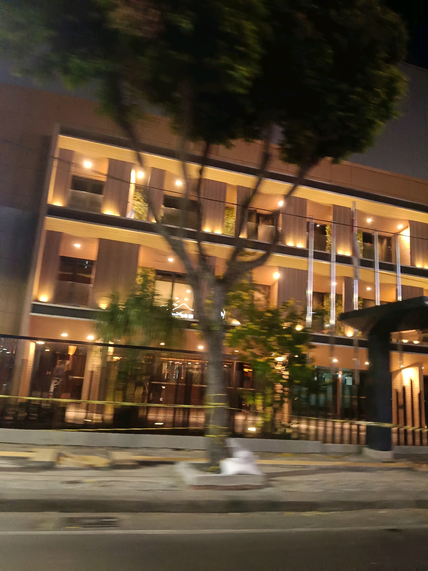 The image shows a building with a modern design at night in Surabaya, Indonesia. The building has multiple floors with large windows and is illuminated from within and by exterior lighting. Trees are visible in front of the building, some of which are partially obscuring the view. Yellow caution tape is stretched across the lower part of the scene, suggesting some kind of barrier or construction. The street in the foreground is visible, and the image appears to have been taken from a moving vehicle due to the motion blur. The overall impression is of an urban environment at night.