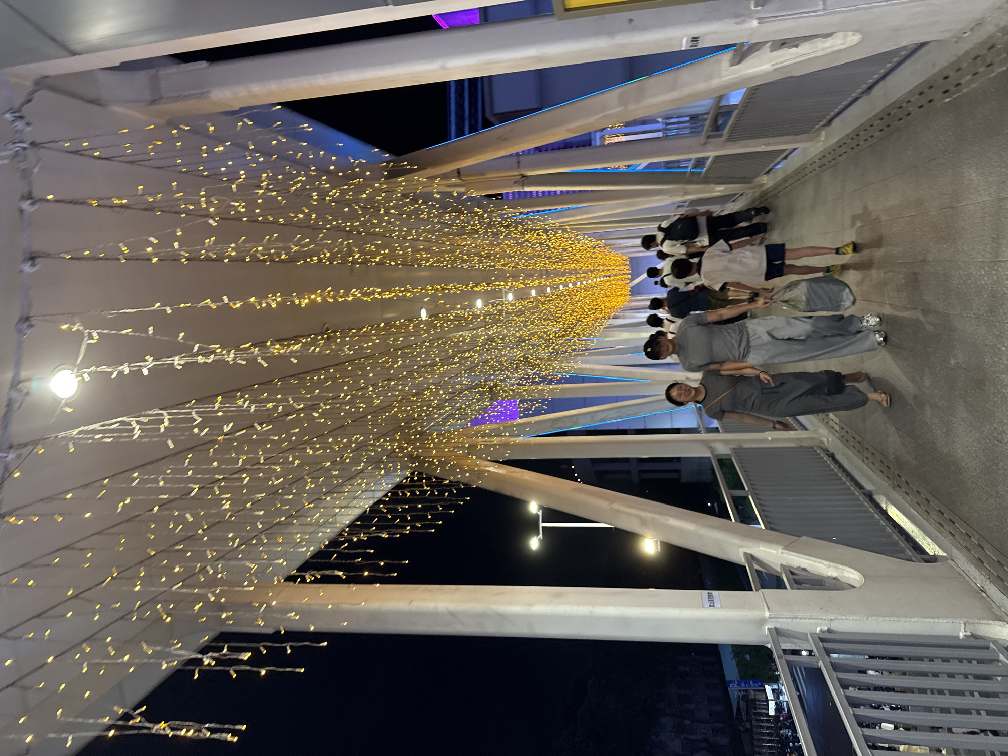 In Tianhe District, China, a pedestrian overpass is illuminated by strings of warm, golden fairy lights hanging from its ceiling, creating a festive atmosphere. The overpass, constructed with white metal beams, spans across a street visible below where motorcycles are parked and streetlights are present. Several people are walking along the overpass, some in pairs and others in groups, moving in the same direction. The individuals are dressed casually, with one person in the foreground carrying a grocery bag. The lighting casts a soft glow, highlighting the texture of the walkway and the railings along the sides. In the background, buildings with blue and purple lighting are visible, suggesting an urban nighttime environment.