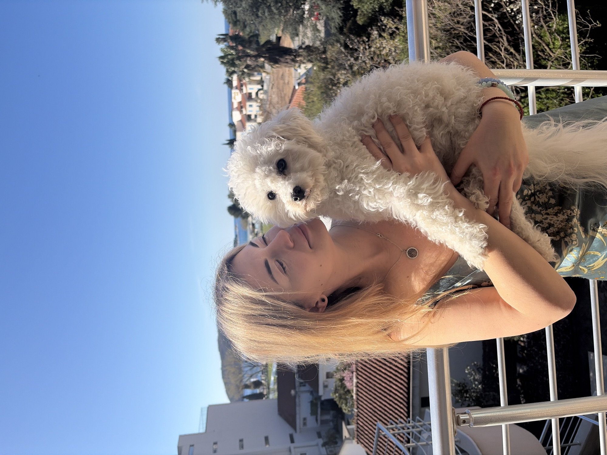 A blonde woman is holding a white Bichon Frise dog on a balcony. The woman has long, light blonde hair and is wearing a necklace and multiple bracelets. She is standing next to a white railing. In the background are buildings with red tile roofs and a clear blue sky. This photo appears to be taken in Budva, Montenegro.