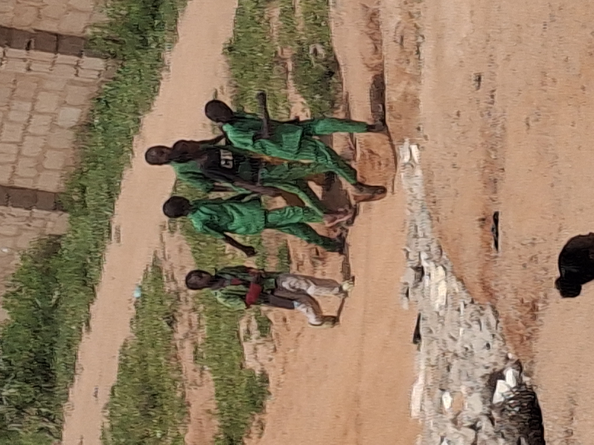 In Tumfure, Nigeria, a group of five young males, likely students, are captured in motion. They are all dressed in green uniforms, with four wearing matching green outfits and one in a black t-shirt with "CM" emblazoned on it, also in green pants. The individuals are walking in a sandy, unpaved area with sparse green vegetation. In the background, a brick wall and more overgrown grass suggest a semi-urban or rural setting. The lighting indicates it is daytime.