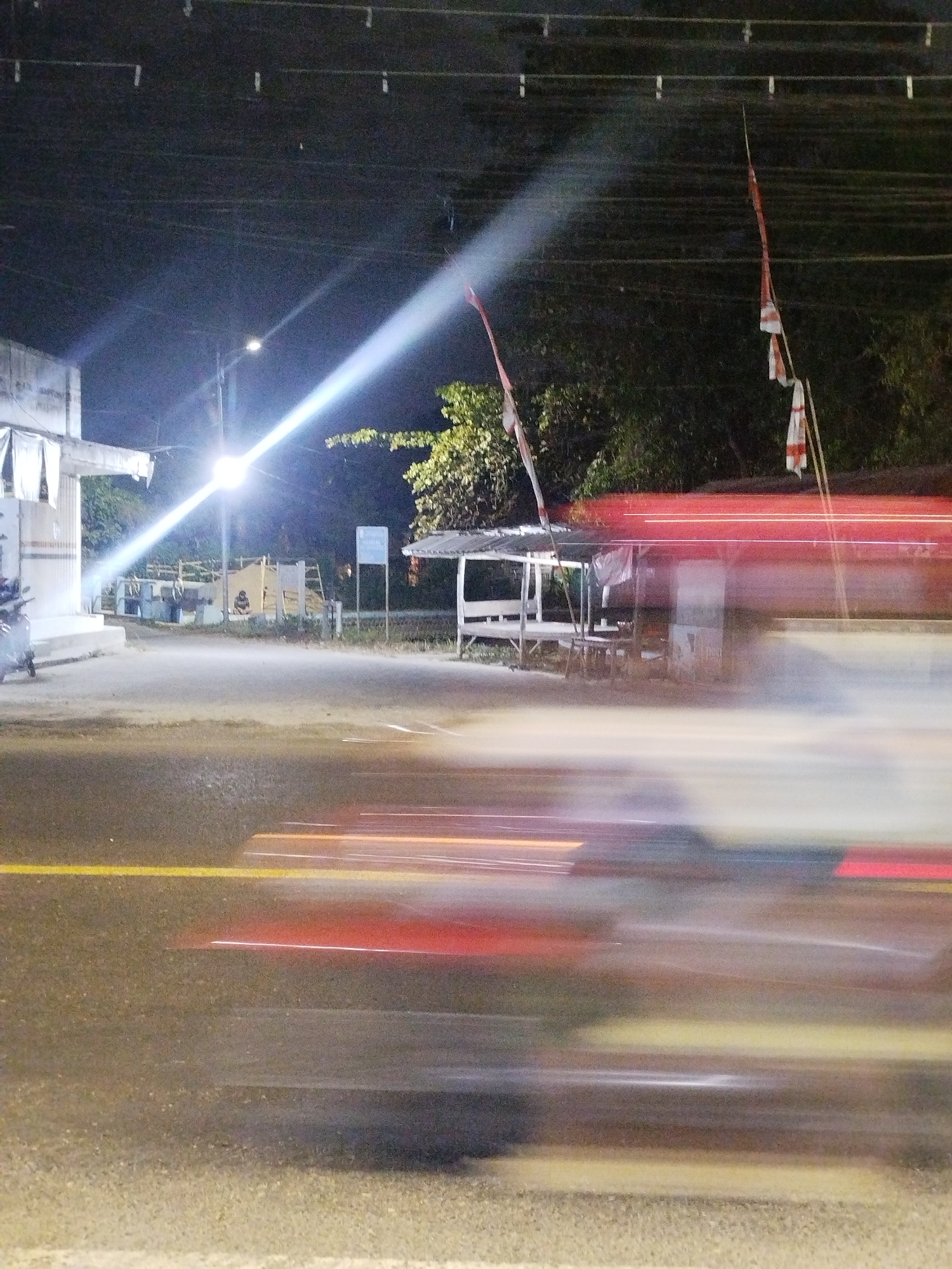 The image captures a night scene on a road in Tonjung, Indonesia, characterized by motion blur due to a long exposure. A bright street lamp illuminates the left side of the frame, casting a strong beam across the road and creating lens flare. On the left, a white building with hanging fabric is visible. Further back, a covered structure with seating and tables is present, with a person sitting near it. Two flags, appearing to be red and white, are displayed on poles. The road itself shows yellow and white lane markings, and the movement of vehicles is indicated by streaks of red and white light. Trees and vegetation line the background.
