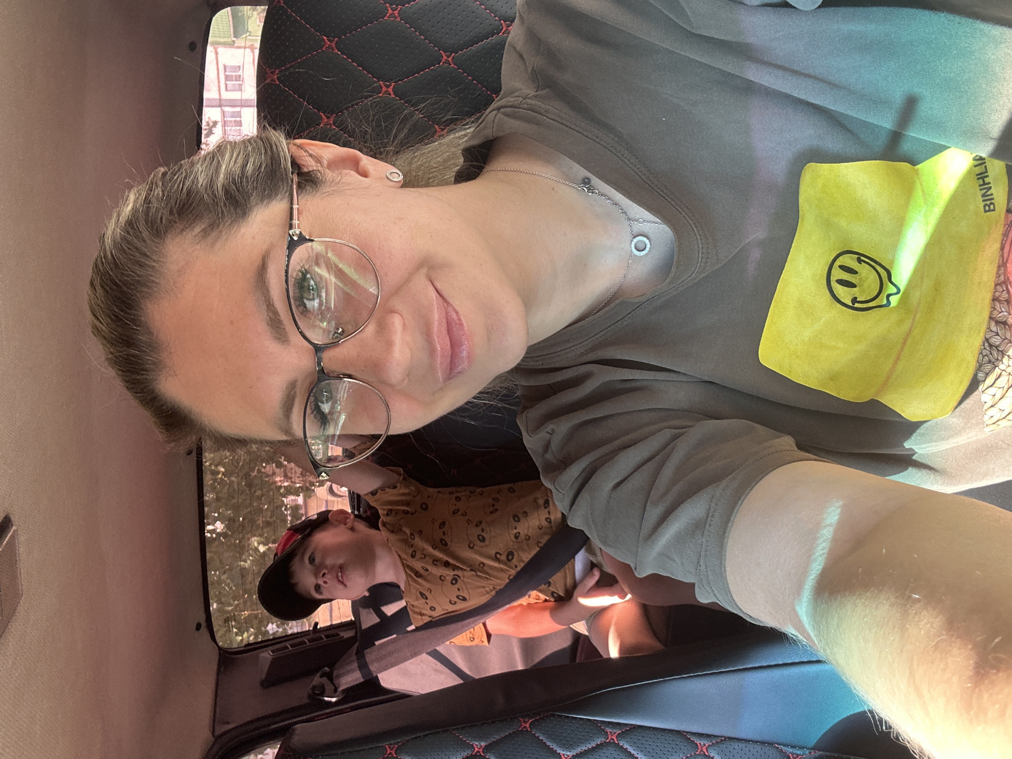 In the car, a woman with blonde hair pulled back and wearing glasses takes a selfie. She has on a green t-shirt with a yellow graphic of a melting smiley face and a silver necklace. To her left, a young boy in a red and black baseball cap and a brown t-shirt with a panda pattern sits in a car seat, looking up. The interior of the car features black seats with red stitching. Sunlight streams into the vehicle, casting shadows. The scene is outdoors, with trees and buildings visible through the car's windows, suggesting a suburban or urban environment likely within Novoaltaysk, Russia, based on the provided location context.