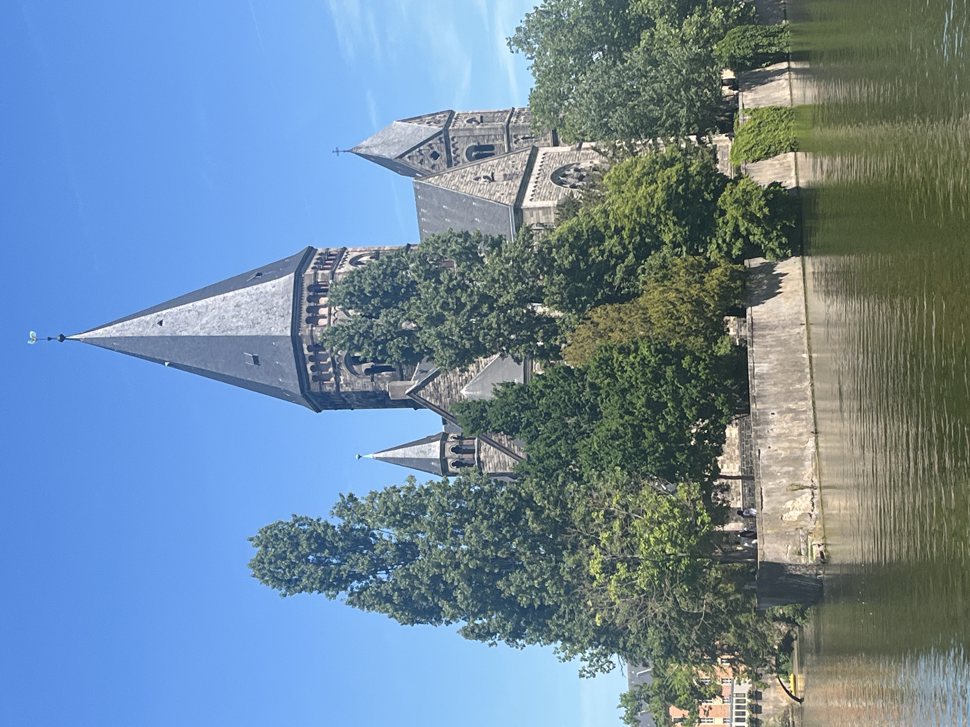 The image captures the Temple Neuf in Metz, France, from a riverside perspective. The neo-Romanesque/Gothic church is surrounded by lush green trees. Part of the bank of the river Moselle is visible. Some people are present on the bank. The sky is bright blue with few clouds.