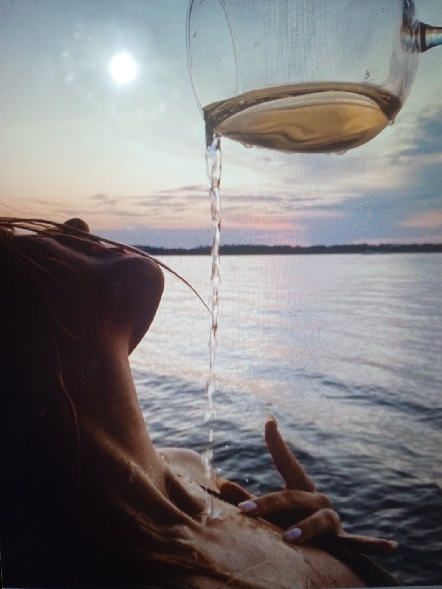 The image shows a person's head and shoulder from behind, with their face turned upwards. A glass of clear liquid is being poured over their head and neck, with the liquid streaming down their skin and into the water. The person is in a body of water, likely a lake or ocean, with a horizon line visible in the background. The sky is a pale blue with some clouds and a bright sun. The lighting suggests it is either sunrise or sunset. There are no visible landmarks or specific features that would indicate a location in Chernivtsi, Ukraine.