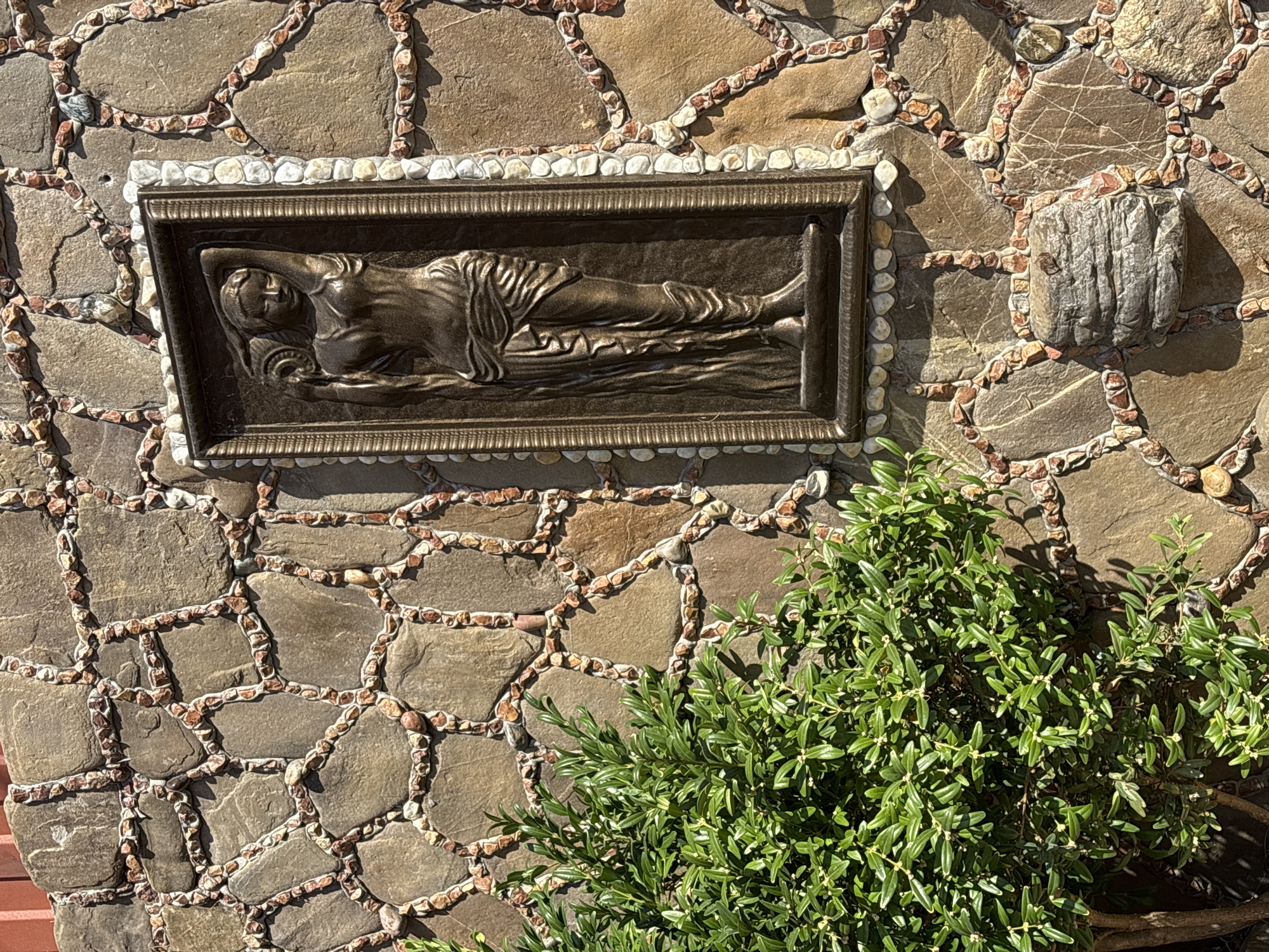 A bronze relief of a female figure is mounted on a stone wall in Razdolnoye, Russia. The wall is constructed with large, irregular brown stones, outlined with smaller, lighter-colored stones and pebbles. The relief, depicting a woman in flowing robes holding a jug, is framed by a decorative border of small, white and gray pebbles. To the left of the relief, a lush green bush with small leaves partially obscures the lower section of the wall. A small, weathered stone ornament is attached to the wall below the relief. The lighting suggests a sunny day, casting shadows on the wall and foliage.