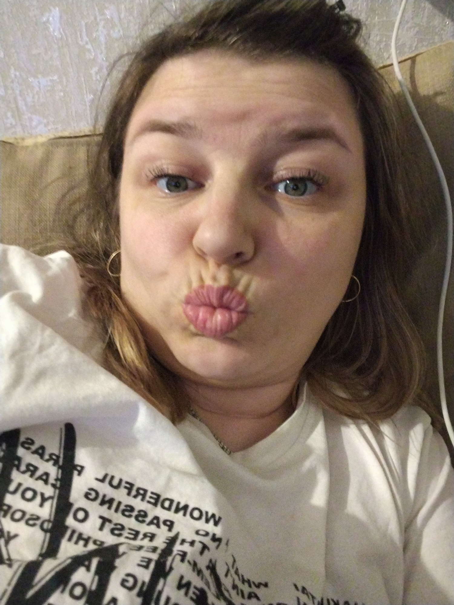 A selfie of a woman with fair skin, likely in her late 20s to mid-30s, with brown hair, making a puckered-lip expression. She has on small gold hoop earrings and a white t-shirt with black text. A thin white cord is visible behind her right shoulder. The background includes a textured wall and a beige-colored surface. The location, Шебунино, Russia is not discernible from the visual information in the image.