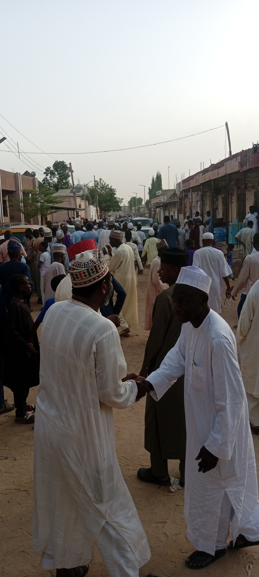 In Sabon Gari West, Nigeria, a large group of people, predominantly men, are gathered in a dusty, open area between buildings. Most individuals are wearing traditional Islamic attire, including white robes and various styles of caps. Two men in the foreground are shaking hands. The buildings in the background appear to be a mix of residential and commercial structures, with some showing signs of age and wear. Overhead, there are utility lines and poles. The sky is overcast, suggesting either early morning or late afternoon.