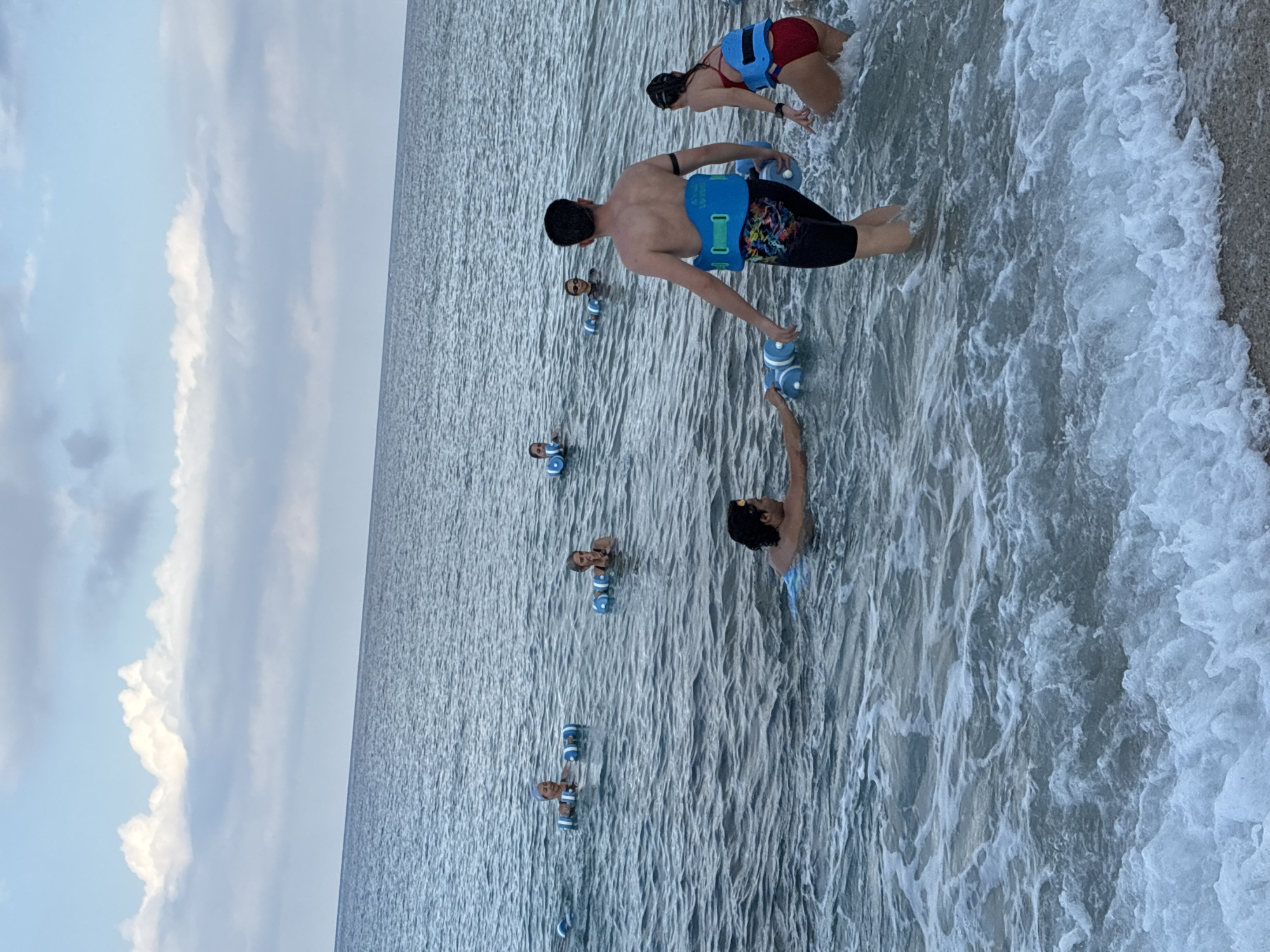 In Alanya, Turkey, a group of people are participating in an aquafitness class in the sea. The water is a moderate depth, with gentle waves lapping at the shore. The sky is overcast with scattered clouds. Most of the participants are wearing flotation belts and holding onto blue flotation devices, suggesting they are engaged in exercises that require buoyancy.
In the foreground, a man with dark hair is partially submerged in the water, facing away from the camera, and appears to be moving. To his right, a woman in a red swimsuit also wears a flotation belt and is partially submerged. Further back, several other individuals are visible, also in the water with flotation devices. Their faces are visible, and some appear to be smiling. The overall scene depicts a recreational activity taking place in a natural marine environment.