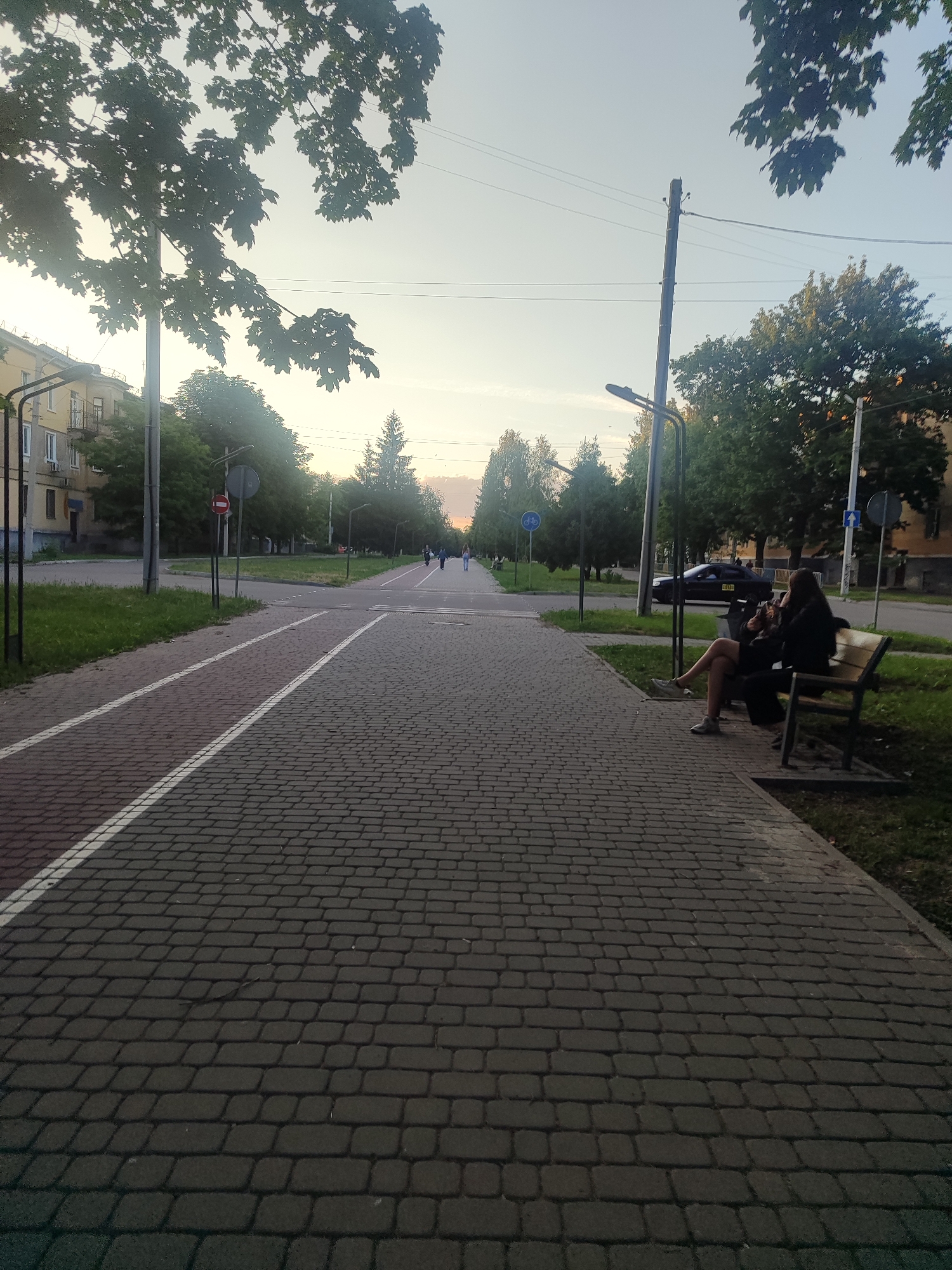 The image depicts a paved pedestrian and bike path in the city of Zhovti Vody, Ukraine, during what appears to be late afternoon or early evening. The path is constructed of grey interlocking pavers, with a dedicated bike lane marked by white lines.
On the right side of the path, a wooden bench is occupied by two individuals. One person, wearing a dark top and shorts, is seated with their legs crossed, holding a mobile phone and appearing to engage with it. Another person, also in dark clothing, is seated beside them.
Further down the path, several other pedestrians are visible in the distance, walking in the same direction. A dark-colored car is parked on the road to the right of the path, and a trash receptacle is situated near the bench.
The environment is characterized by mature trees lining the path, with some foliage casting shadows. Streetlights are present along the route, and buildings are visible on either side of the street. The sky is a pale, clear hue, suggesting calm weather conditions. The overall scene conveys a sense of urban tranquility.
