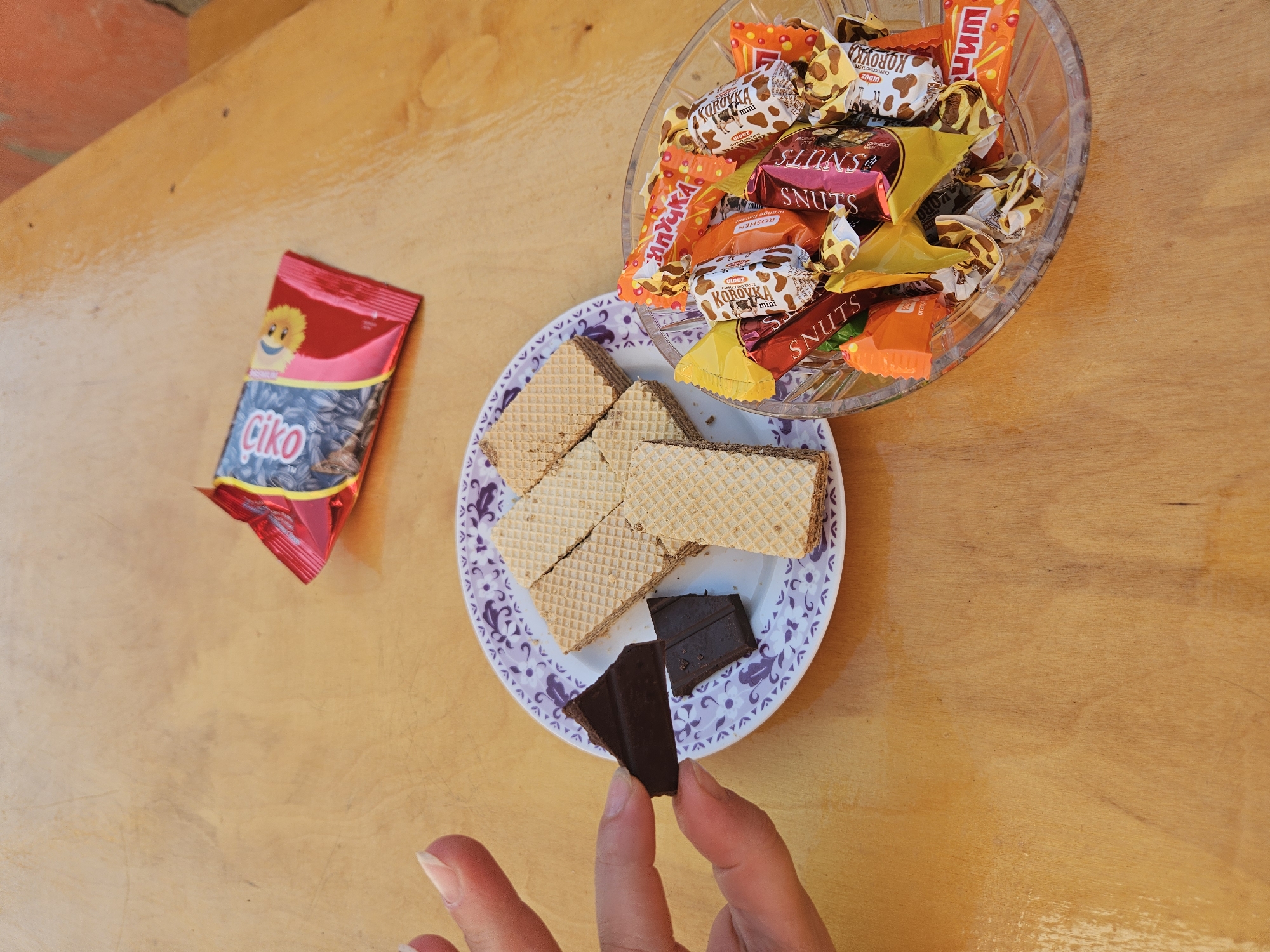 A close-up, eye-level shot captures a wooden table surface with various snacks. In the upper right, a red and pink foil packet of "ako" brand sunflower seeds lies open, revealing dark seeds within. To the left, a white plate with a purple decorative rim holds several rectangular wafer cookies. In the foreground, a person's hand is visible on the left, holding two dark chocolate pieces. To the right of the plate, a clear glass bowl is filled with an assortment of individually wrapped candies in various colors and wrappers, including "SNUTS," "Korovka mini," and other unidentifiable brands. The lighting suggests an outdoor setting, likely during daylight hours, with the focus on the collection of snacks.