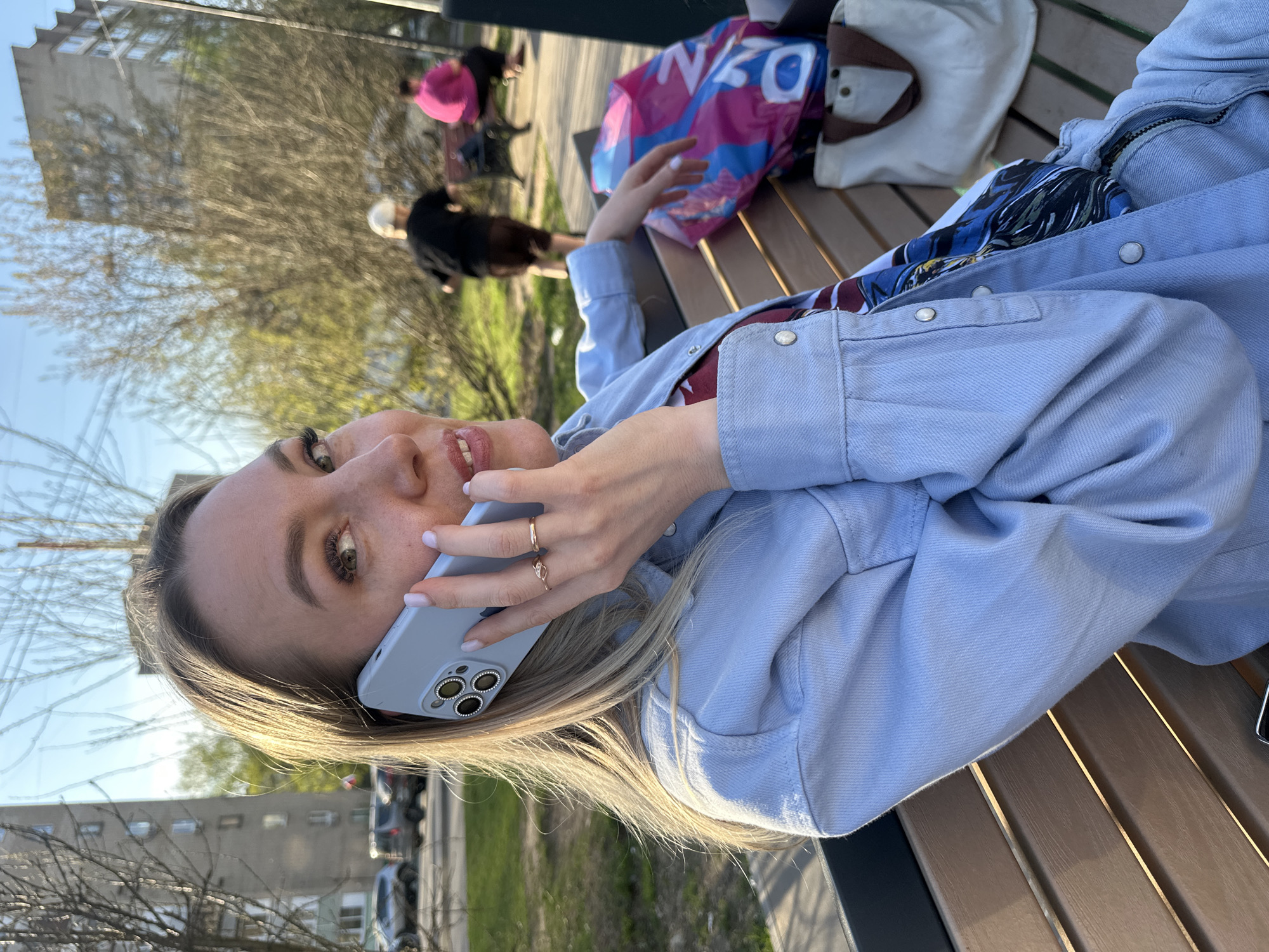 A woman is lying on a park bench talking on a smartphone. She is wearing a light blue denim jacket and has blonde hair. A pink and blue shopping bag is on the bench next to her. In the background, there are other people on benches and trees. Buildings can be seen in the far background. The scene appears to be in the city of Tomilino, Russia.
