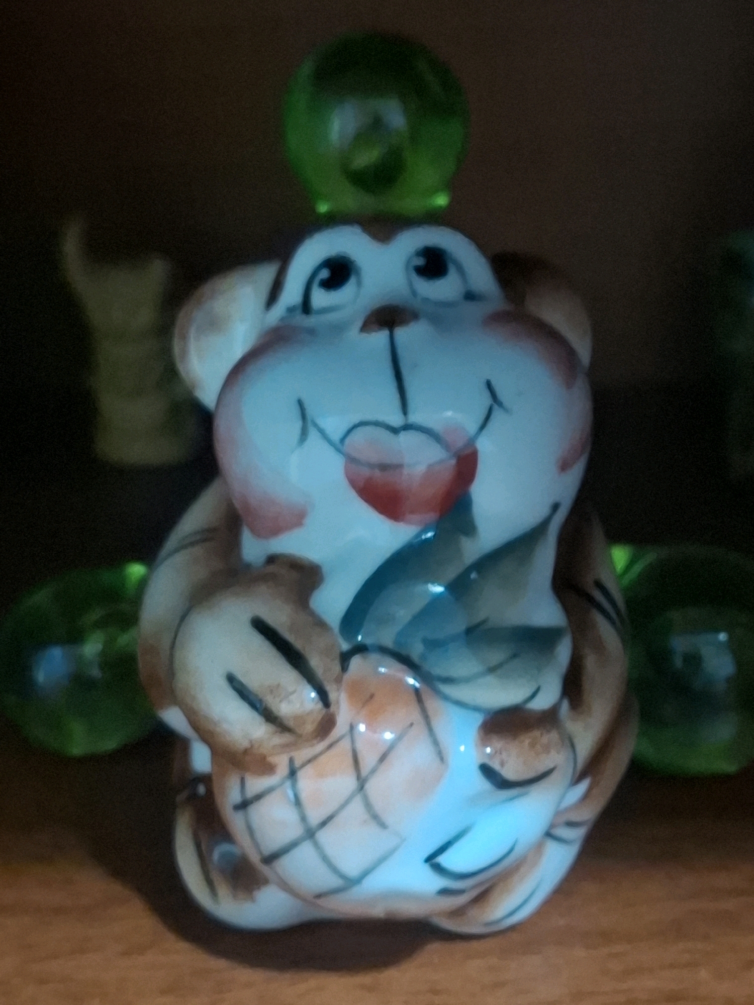 A ceramic figurine of a monkey sits on a wooden surface. The monkey is depicted with large, round eyes, a smiling mouth, and rosy cheeks. It is holding an orange object, possibly a fruit or a basket, with both hands. The figurine is decorated with brown and white colors, with accents of red and blue. A green sphere rests on top of the monkey's head, and two other green spheres are visible on either side of the figurine. The background is dark and out of focus, suggesting an indoor setting. The overall lighting is dim, with some areas illuminated by a bluish light. There is no information available to determine the location in the city of Almaty, Kazakhstan.