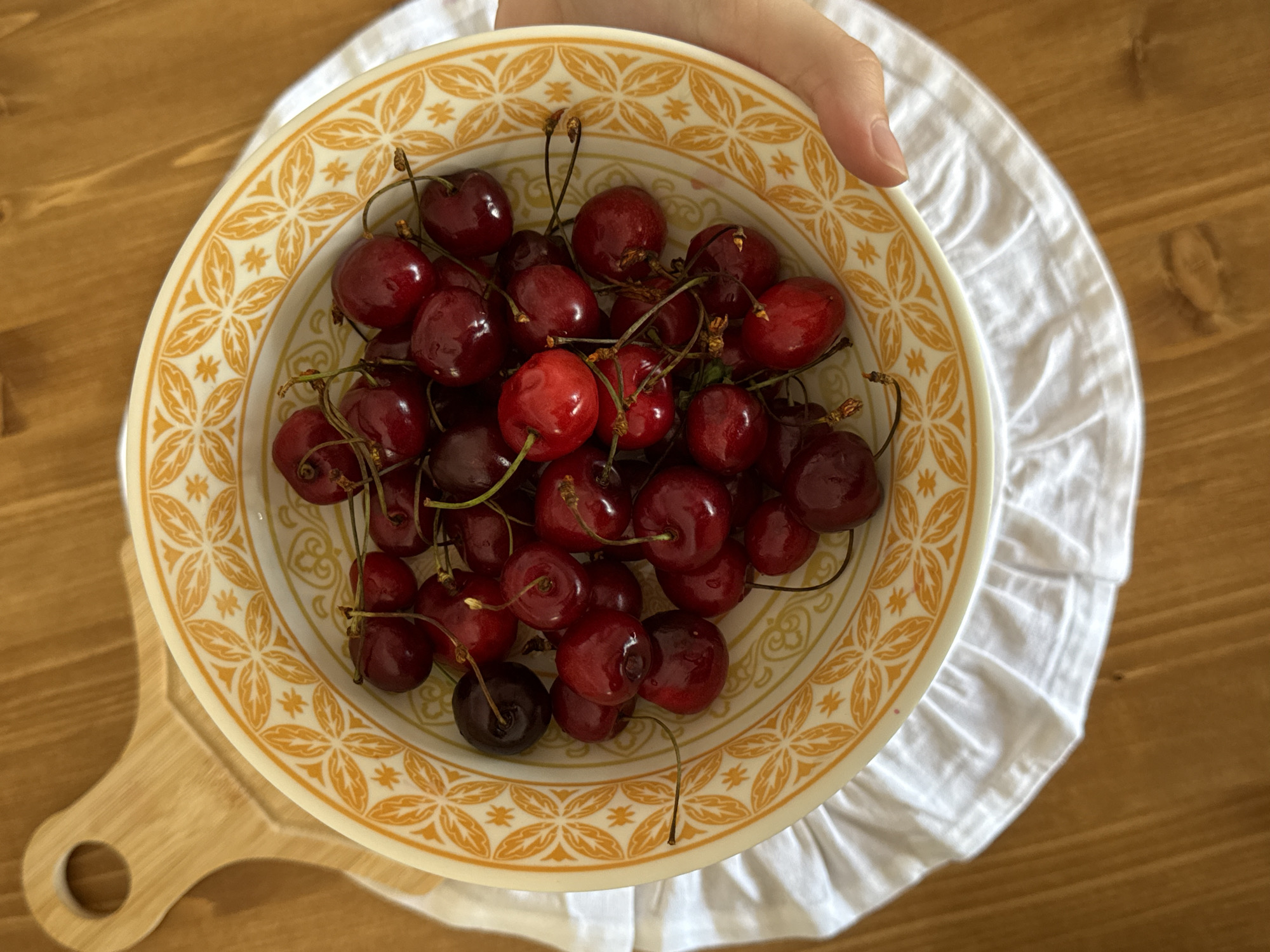 A close-up, top-down shot shows a person's hand holding a yellow-rimmed bowl filled with ripe cherries. The bowl features a decorative pattern of orange floral motifs. The cherries are a deep red color and have stems attached. They are piled in the bowl, with some slightly darker, almost black cherries mixed in. The bowl sits on a white ruffled cloth, which is placed on a wooden surface. To the left of the bowl, a portion of a wooden cutting board with a circular handle is visible. The lighting suggests it is daytime, and the scene appears to be indoors. No visible text is present in the image. The overall impression is one of freshness and natural abundance.