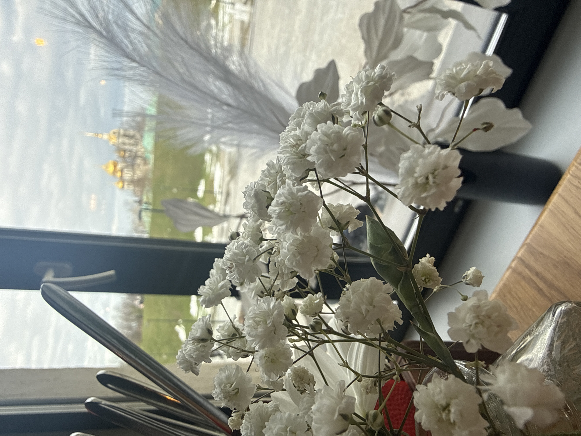 The image shows a bouquet of white gypsophila flowers in a vase, likely placed on a table near a window. Through the window, a blurry view of the Assumption Cathedral in Dmitrov, Russia is visible. Also seen are some silverware, a black rectangular object, and a brown wooden surface. The scene suggests an indoor setting, possibly a restaurant or cafe, with the flowers serving as decoration. The light suggests daytime.