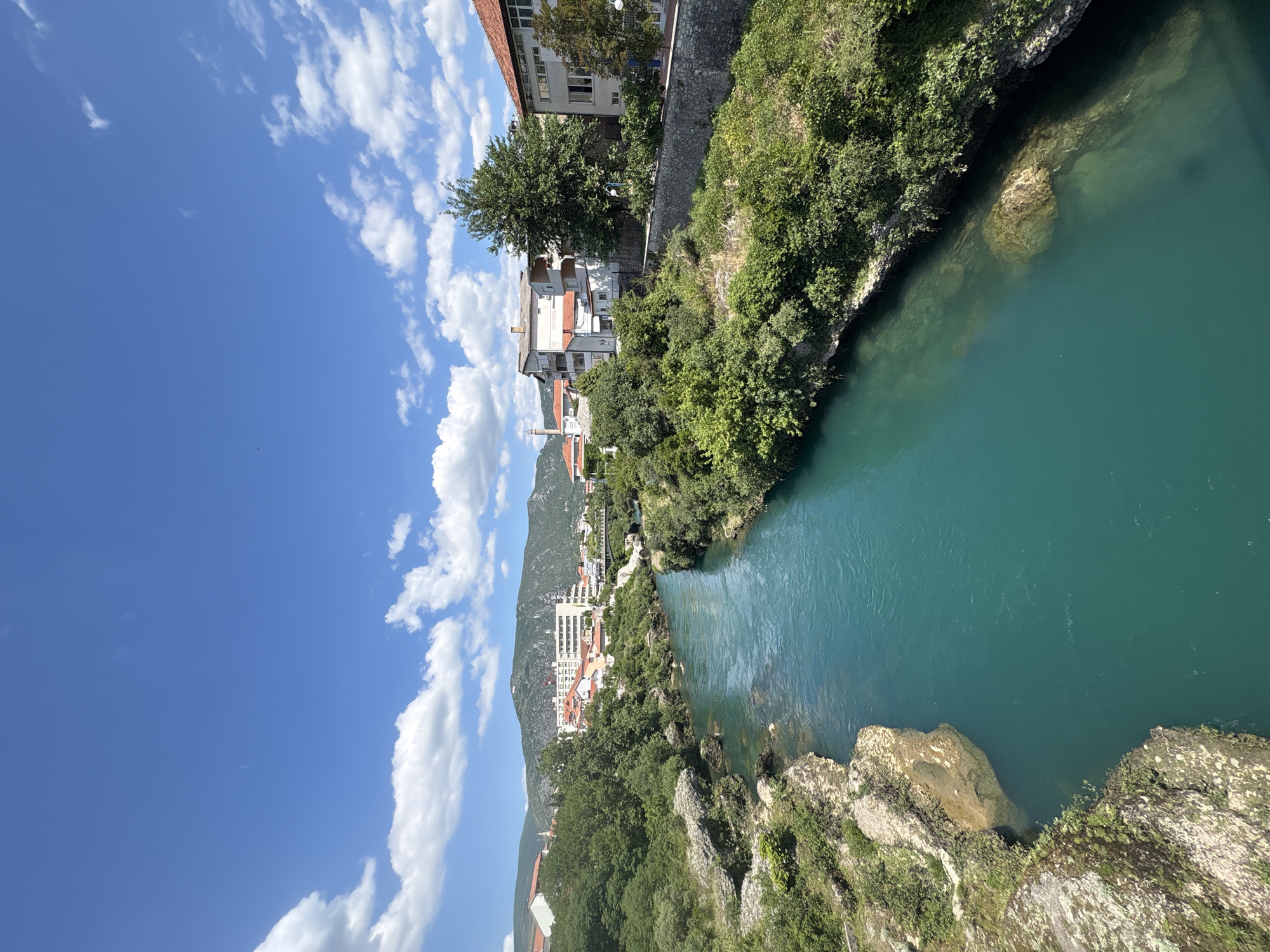 The image shows the Neretva River as it passes through Mostar, Bosnia and Herzegovina. The river, a striking turquoise color, is flanked by rocky, vegetation-covered banks. Buildings can be seen rising along the riverbank on one side, against the backdrop of a mountain. The sky is blue with scattered white clouds.