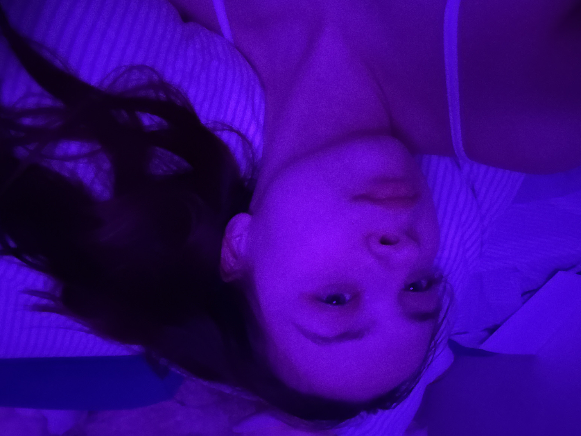 A young woman lies on a striped bed, illuminated by blue light. She faces the camera with a neutral expression. Her dark hair falls beside her head. She wears a light-colored tank top. The location of the photo is unknown and there is no information available to indicate it was taken in Прикамский, Russia.