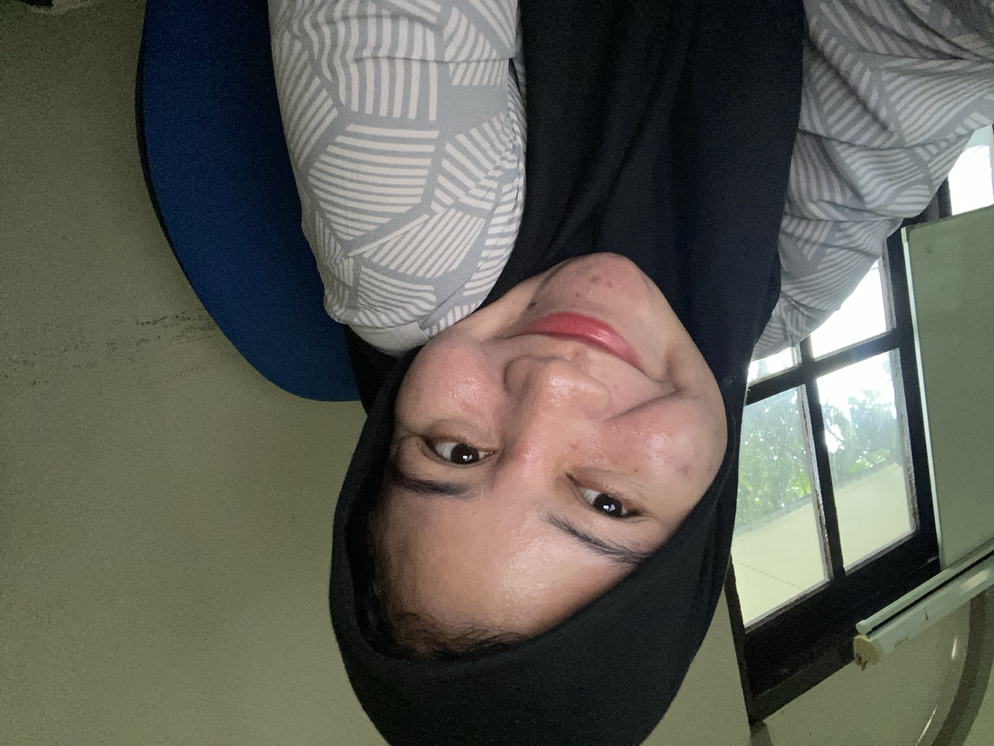 The image is a selfie of a woman wearing a black hijab and a gray and white patterned shirt. She is smiling and looking towards the camera. The background appears to be indoors, with a window visible on the left side of the frame, showing trees and daylight outside. A portion of a blue chair is also visible on the right. The lighting suggests it is daytime, and the overall setting seems to be a residential or office space. There are no specific visual cues that tie this image to Banjarbaru, Indonesia, as the location is not explicitly indicated in the visual content.