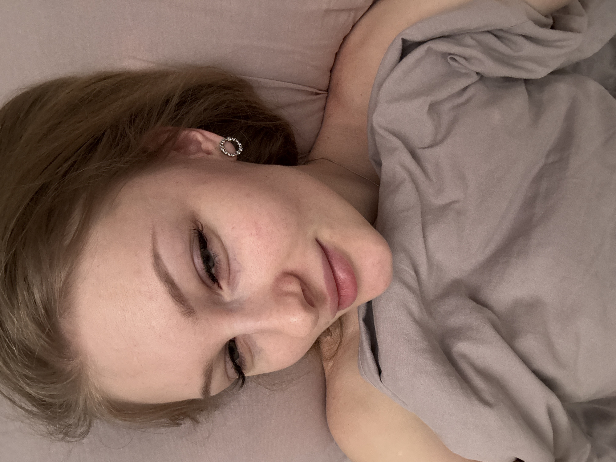 A fair-skinned female lies on a lavender-colored pillow and is partially covered by a gray sheet. Her head is tilted to the right and her eyes are closed. She has brown hair and is wearing a circular, possibly rhinestone, earring in her left ear. No indication of the image's location in Perm, Russia is discernible.