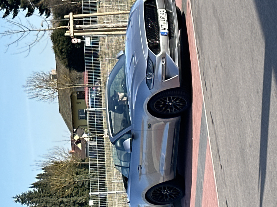 A silver Ford Mustang convertible, model year 2018 or newer, with its top retracted, is positioned on a paved area displaying red and dark gray markings. A driver, identifiable by a dark baseball cap, occupies the driver's seat. The vehicle bears a German license plate, "WT IR 41," which signifies registration within the Waldshut-Tiengen district, situating the scene in Jestetten, Germany. The Mustang features a "5.0" badge on its side panel and black multi-spoke wheels. In the background, behind a metal fence, are several residential buildings, including a light yellow house with a dark gabled roof. Bare deciduous trees and evergreen trees are visible. The sky is clear blue. A wooden utility pole, equipped with a small circular sign, stands on the right side of the frame. Shadows are cast on the foreground pavement, indicating direct sunlight.