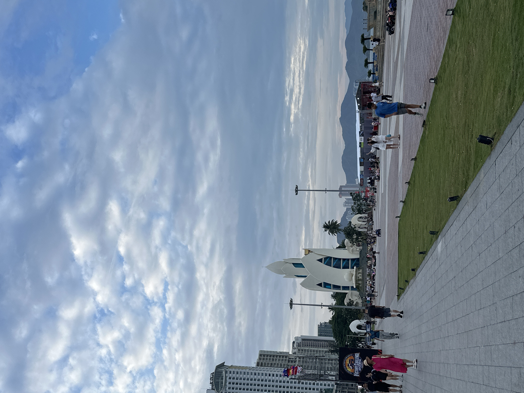 This image captures a vibrant outdoor scene in Nha Trang Ward, Vietnam, featuring a spacious public plaza flanked by a strip of green grass on the right. The sky above is largely covered with varying shades of grey and white clouds, with glimpses of blue sky appearing in certain areas, suggesting an overcast day.
Numerous people are scattered across the paved areas, walking and congregating. In the mid-ground, a man in a blue t-shirt and grey shorts walks with purpose, while further back, a crowd has gathered around a stage or event structure. Closer to the foreground, a woman in a bright pink dress and a man in a black t-shirt are seen walking along the grey paved pathway. Small, dark ground spotlights are embedded along the edge of the grassy area.
A prominent architectural feature is a unique, white building with a distinctive, somewhat organic or lotus-like design, featuring blue-tinted windows. This structure is surrounded by trees, including palm trees, and modern high-rise buildings. In the distant background, a range of mountains rises against the horizon.
A large black banner or sign is partially visible in the bottom left corner, clearly displaying the text "VICTORY CHALLENGE CUP" and "2023", indicating a specific event is likely taking place. The overall impression is that of a public event or celebration, with people enjoying the outdoor space. The time of day appears to be daytime, possibly late afternoon, given the ambient light despite the cloud cover.