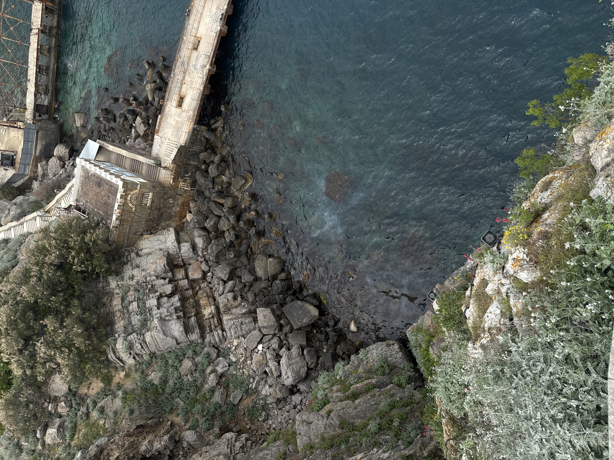 This is an aerial view taken in Гаспринский поселковый совет, Ukraine, showcasing a rugged coastline with a mix of natural and man-made features.
The image features:
1. **Water**: A body of dark water with visible ripples and some transparency, allowing glimpses of the underwater rock formations.
2. **Rocks and Cliffs**: A combination of large, angular rocks and layered cliffs, covered with vegetation.
3. **Vegetation**: Various types of plants, including bushy vegetation, grasses, and small flowers.
4. **Infrastructure**: Segments of what appear to be old piers or docks.
5. **Buildings**: There is a small building complex with staircases and other architectural features.
The photo offers a direct, overhead perspective. There are no people visible in the image. The overall scene conveys a sense of the coexistence of nature and human influence in this coastal region.