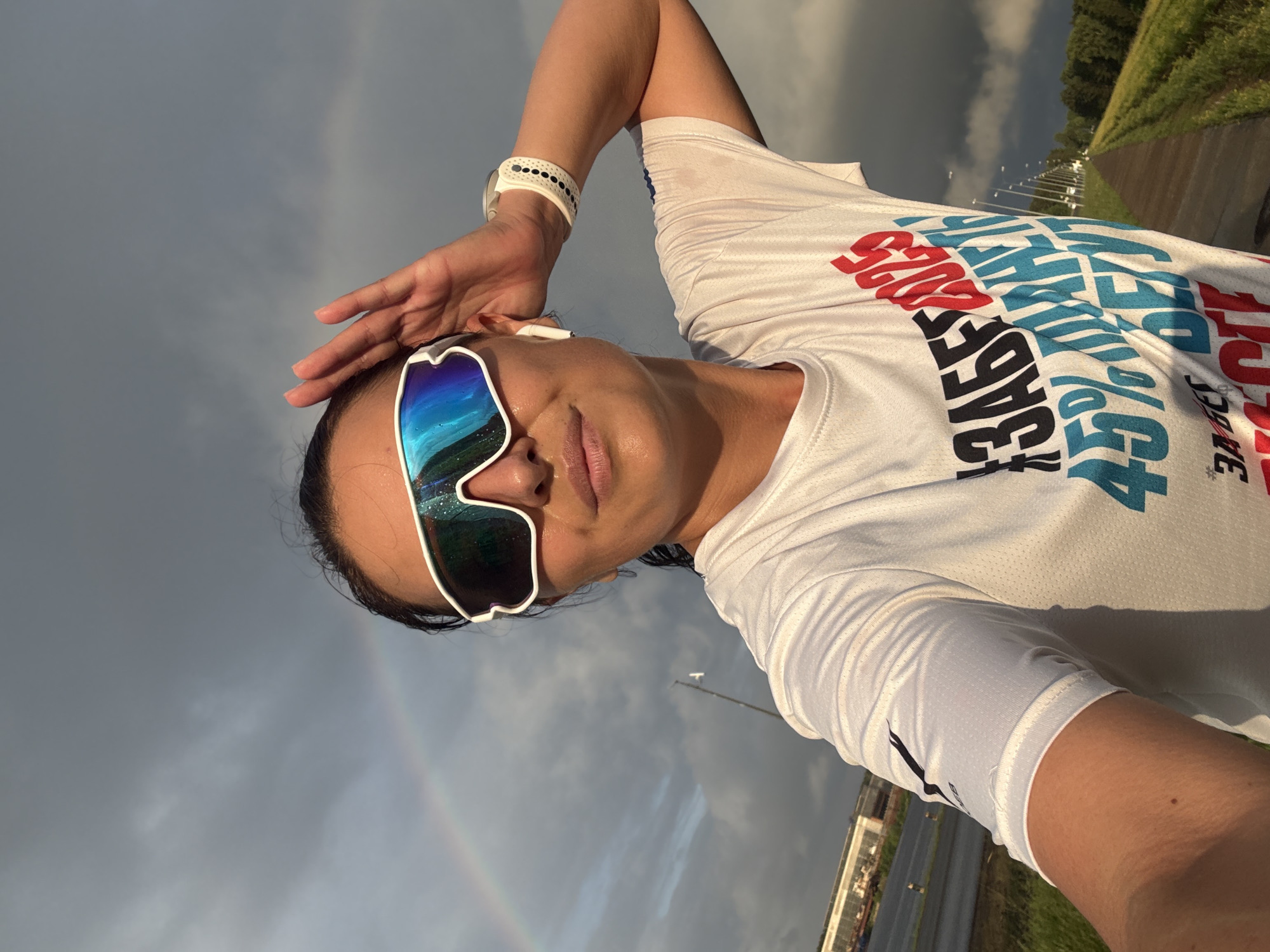 A woman with dark hair, wearing athletic sunglasses and a white t-shirt with blue and red lettering, takes a selfie outdoors. She is holding her right hand up to her head. A faint rainbow is visible in the cloudy sky behind her. The background shows a road, some buildings, and trees, suggesting an urban or suburban setting. The t-shirt has Cyrillic text on it, including "ЗАБЕГ" (run) and "2025". The scene appears to be a park or a path near an industrial area. The lighting suggests it is either early morning or late afternoon, possibly after a rain shower.