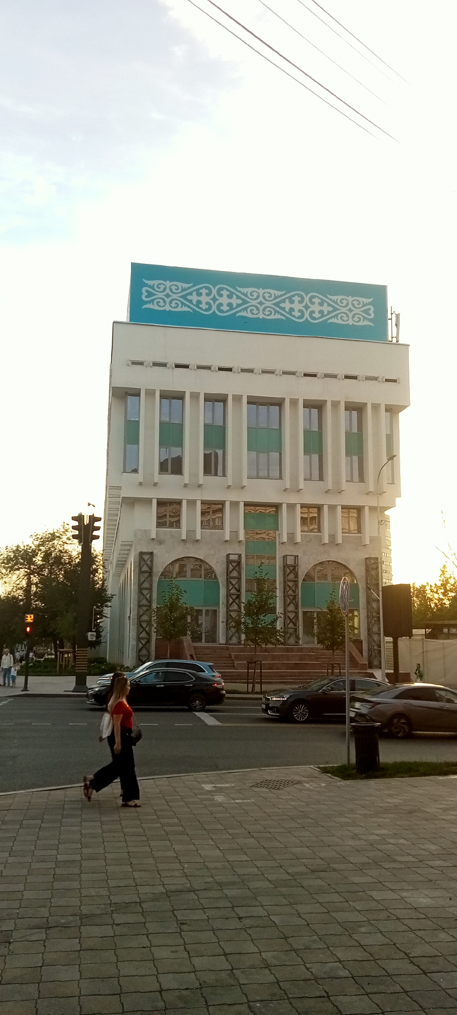 The image depicts a street scene in Almaty, Kazakhstan, with a multi-story building as the central focus. The building features a modern architectural style with a white facade and teal decorative elements at the top, including an intricate pattern. The lower levels of the building have arched entrances and large windows with teal accents. In front of the building, there is a wide street with several cars, including a black sedan and a dark blue SUV. A pedestrian, a woman wearing a red top and dark pants, is walking on the sidewalk. Traffic lights and trees are visible along the street. The sky is bright and appears to be late afternoon or early evening, casting a warm light on the scene.
