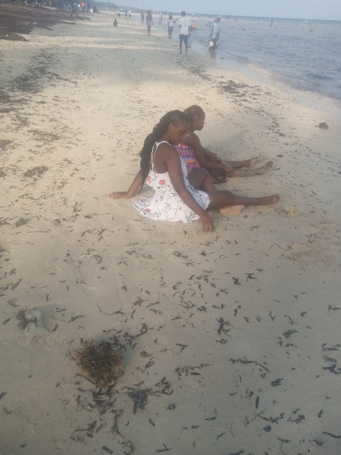 The image displays a sandy beach scene in Shanzu ward, Kenya. In the foreground, two individuals are seated on the light-colored sand, facing towards the right side of the frame. The individual closest to the camera wears a white sleeveless dress with a scattered dark pattern and has long, dark braided hair. Her arms are extended onto the sand, and her legs are stretched forward. Behind her, a second individual, wearing a patterned garment and possessing lighter-colored braided hair, is similarly seated. The sand directly in front of and around these two individuals is littered with scattered dark organic debris, primarily appearing to be seaweed. In the midground, numerous individuals are visible walking along the sandy beach near the waterline. The background consists of a calm, light blue ocean, where additional people are observed wading or swimming. The sky above the horizon appears overcast or hazy.