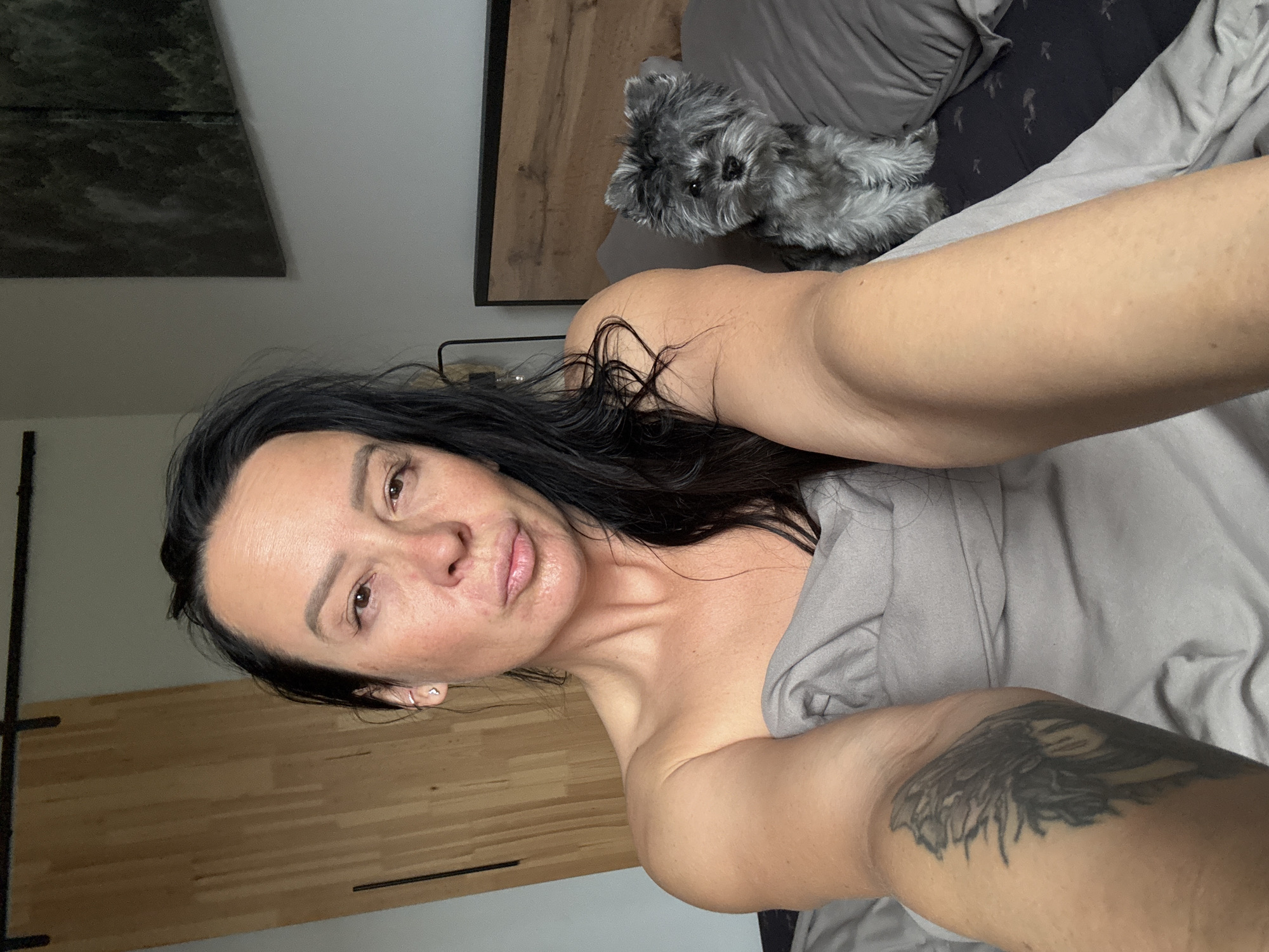 A selfie taken indoors, likely in a bedroom. The subject is a woman with dark hair and a visible tattoo on her arm, partially covered by a gray blanket or sheet. She is lying in bed. A small, gray, fluffy dog is sitting on the bed near the headboard. A picture hangs on the wall. The location is not recognizable as Chaikovsky, Russia.