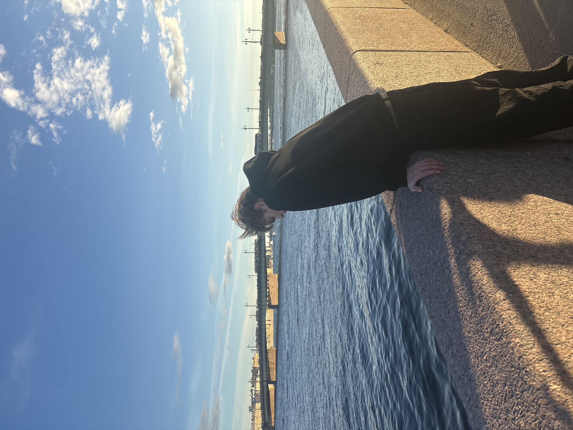A young person wearing a black hoodie and pants leans over a stone embankment, looking down at a body of water. The embankment is made of rough, textured stone blocks that are illuminated by warm, low-angle sunlight, casting long shadows. The water is dark blue with ripples, suggesting a slight breeze. In the background, a long bridge spans the water, with buildings and what appear to be trams or trains visible on it. The sky is a clear blue with scattered white clouds, illuminated by the setting sun. The scene suggests a contemplative moment by the water in an urban setting. The location appears to be a waterfront area in Gorbunki, Russia, likely near a bridge and embankment typical of Russian cities.