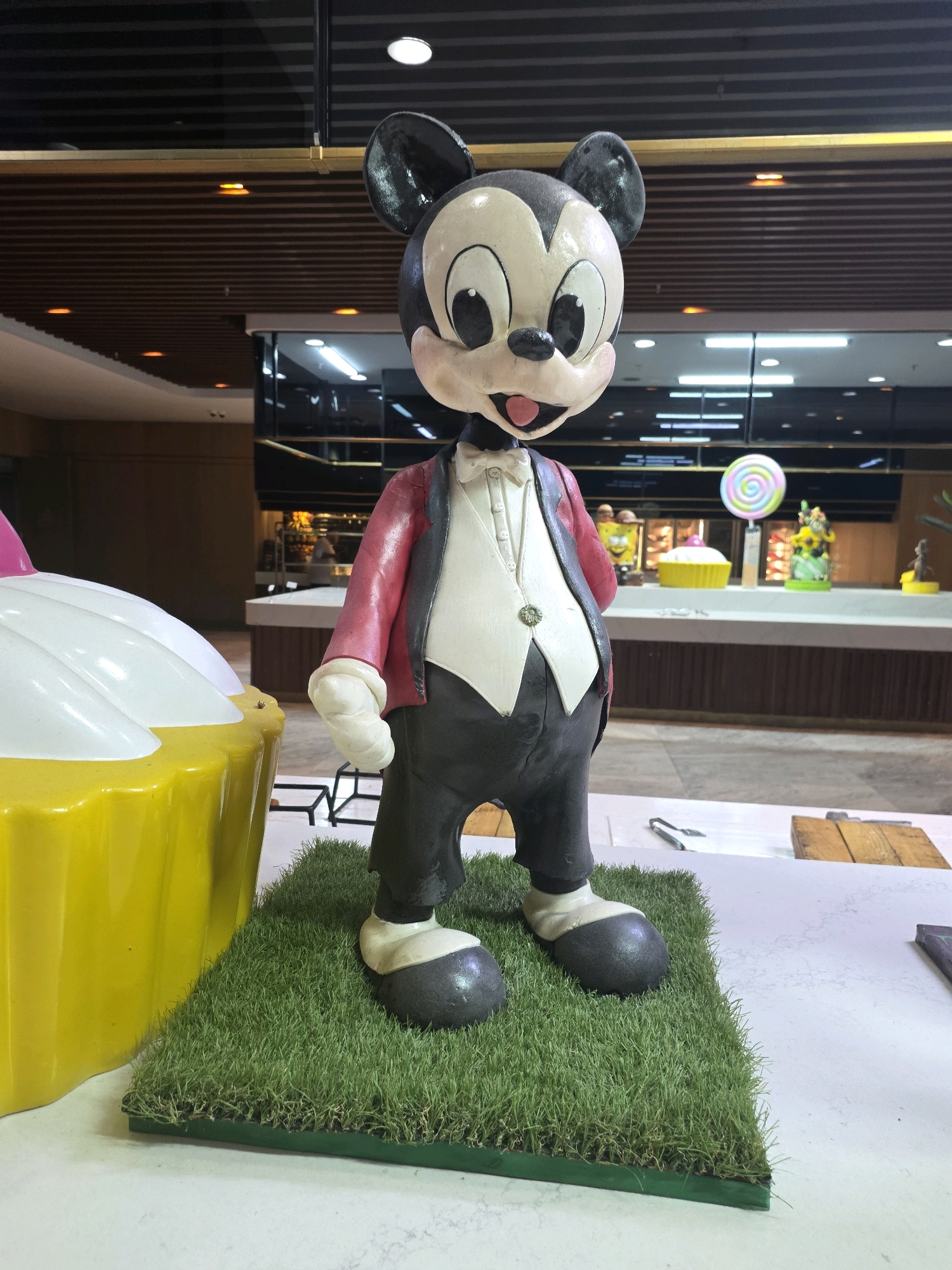 The image shows a statue of Mickey Mouse, dressed in a red jacket, white vest, and black pants, standing on a patch of artificial grass. To the left of the statue is a large, yellow, cupcake-shaped decoration with white frosting. In the background, there appears to be a store or market with various colorful displays, including a lollipop and more cupcake decorations. The ceiling has wooden slats and overhead lighting. The scene is indoors, likely in a public area.